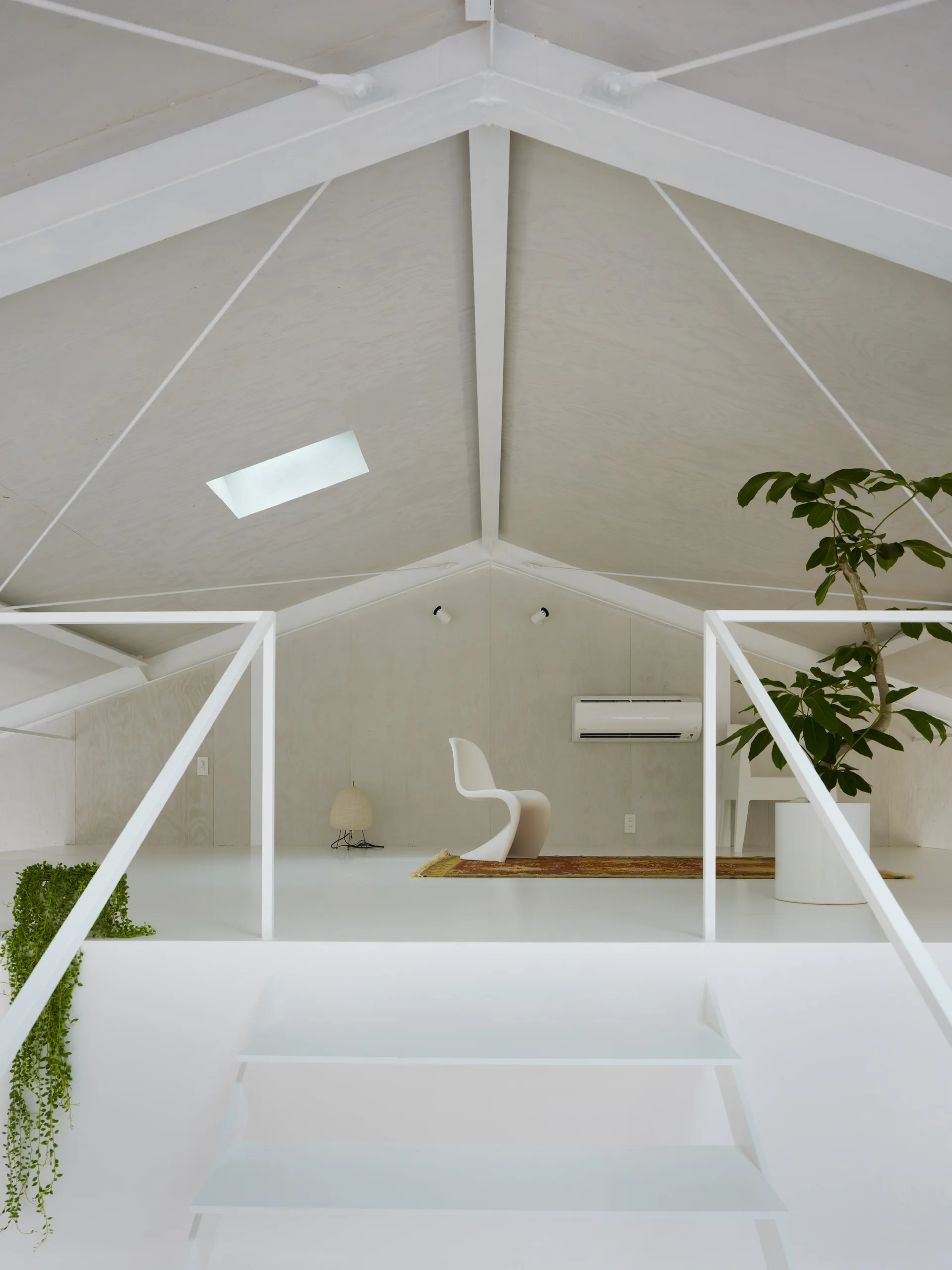 A bright attic loft features white stairs, a vaulted ceiling with a skylight, and a modern white chair on a patterned rug against plywood walls.
