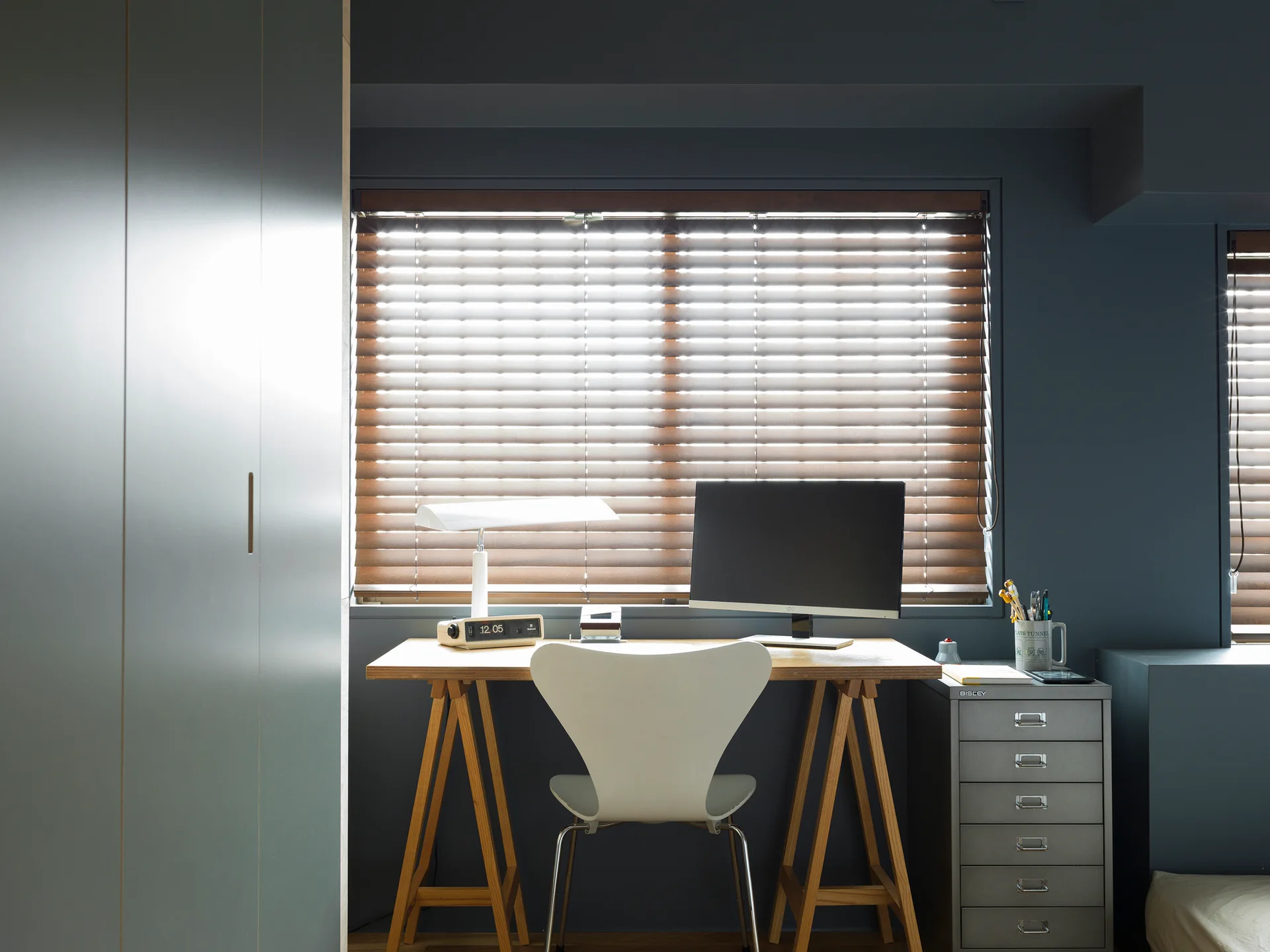 A home office features a wooden trestle desk, white modern chair, and metal file cabinet set against dark charcoal walls under a window with wooden blinds.