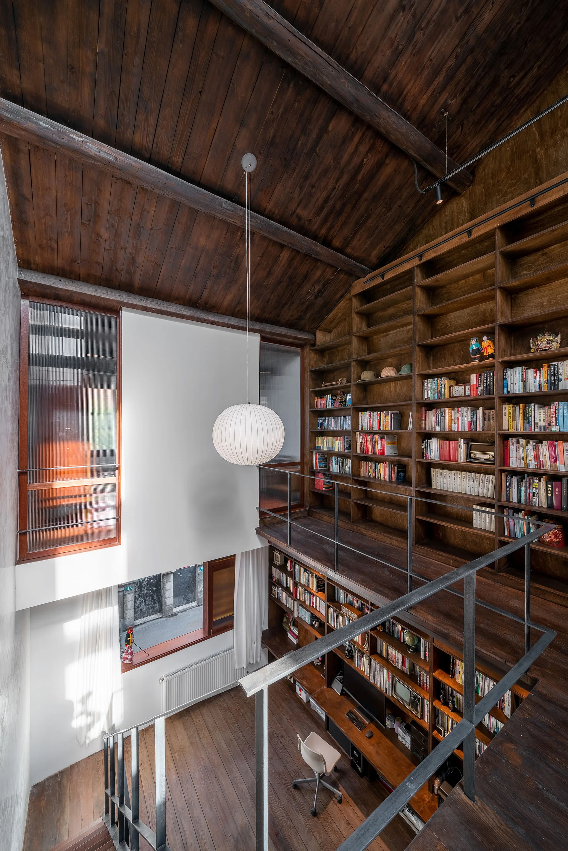 A multi-level home library with tall dark wood bookshelves, a wooden plank vaulted ceiling, a mezzanine level, and a white hanging pendant lamp.