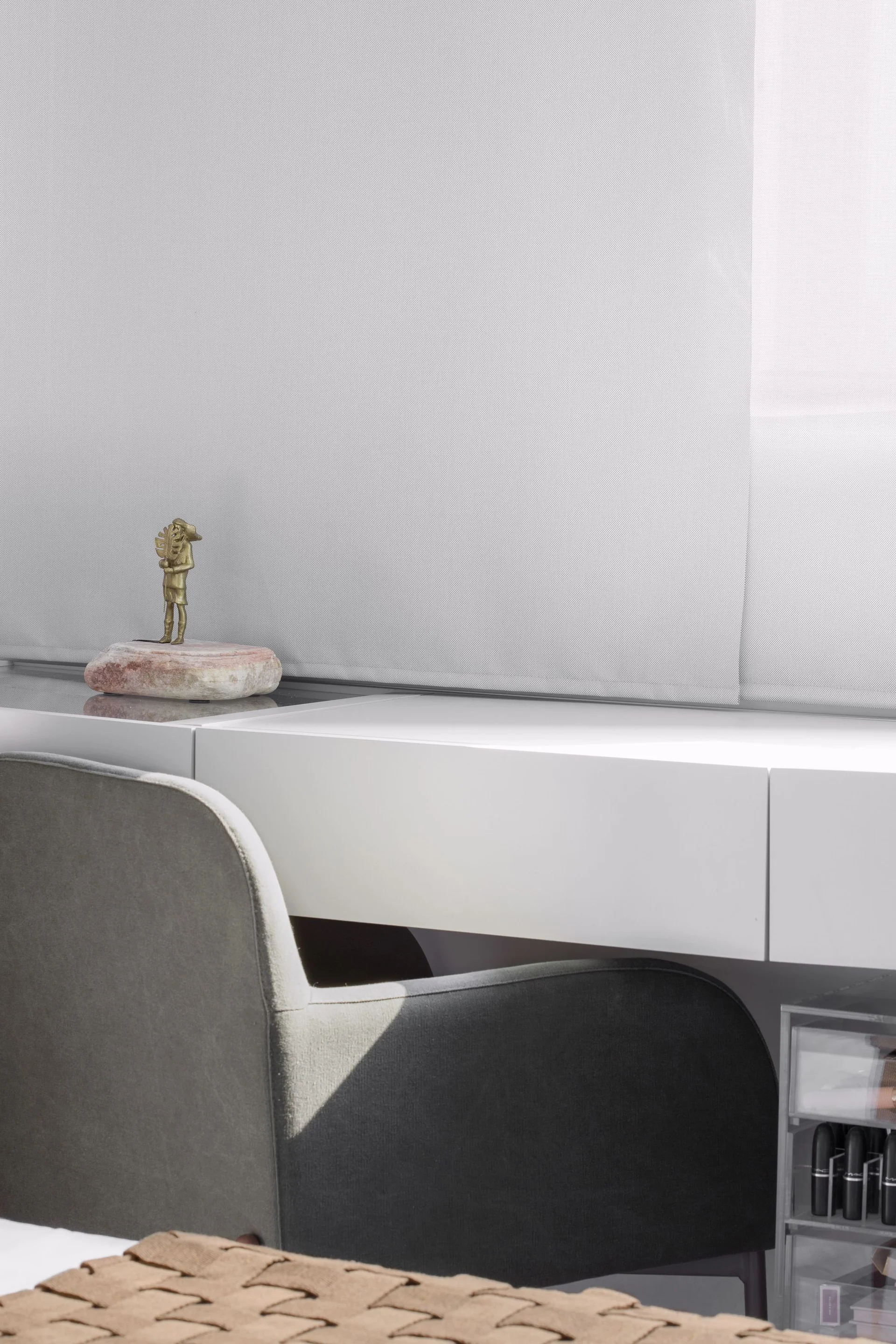 A white minimalist desk is paired with a gray upholstered armchair and styled with a small brass sculpture on a stone base.