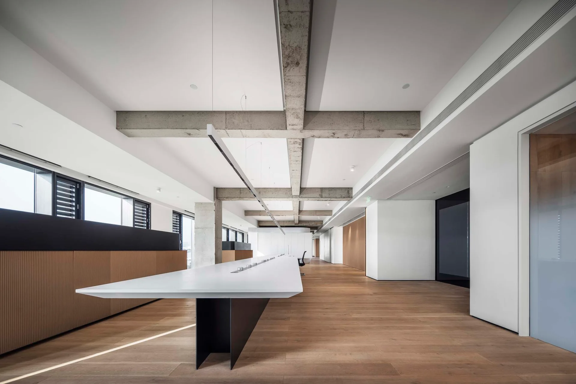 A long white desk sits on wood flooring in a spacious, minimalist office with exposed concrete ceiling beams and slatted wall panels.