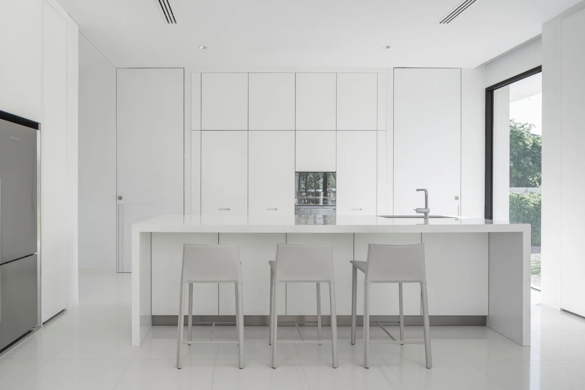 A minimalist white kitchen features handleless floor-to-ceiling cabinets, a large central island with three grey stools, and shiny tiled floors.