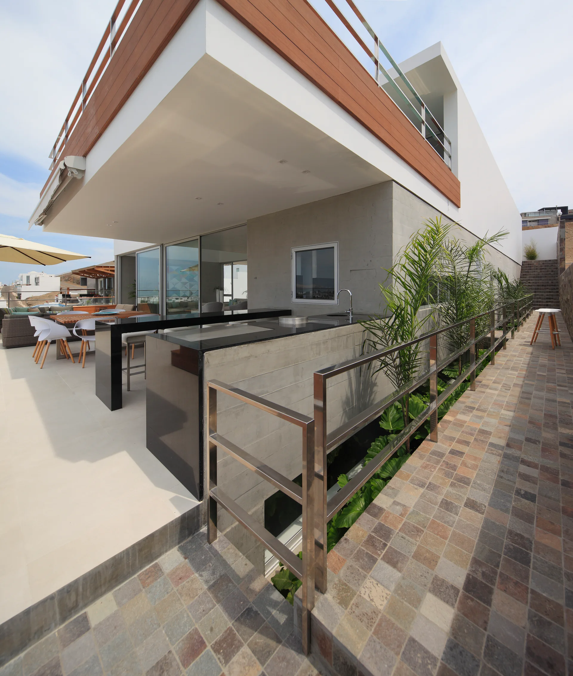 A modern outdoor terrace features a board-formed concrete kitchen island with a black countertop, metal railings, and a tiled walkway.