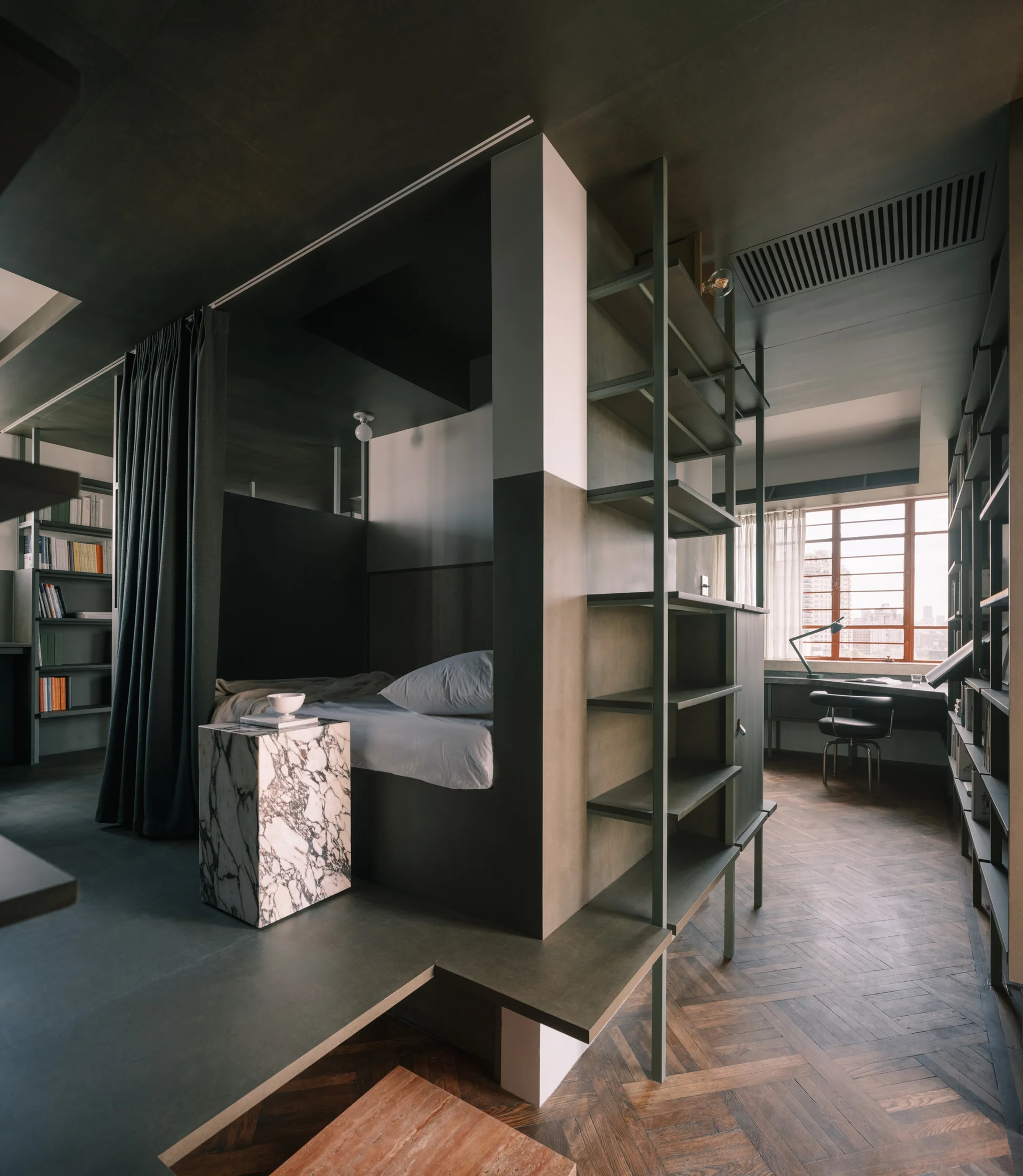 A dark charcoal modern bedroom featuring an integrated metal shelving unit, a platform bed, a veined marble side table, and herringbone flooring.