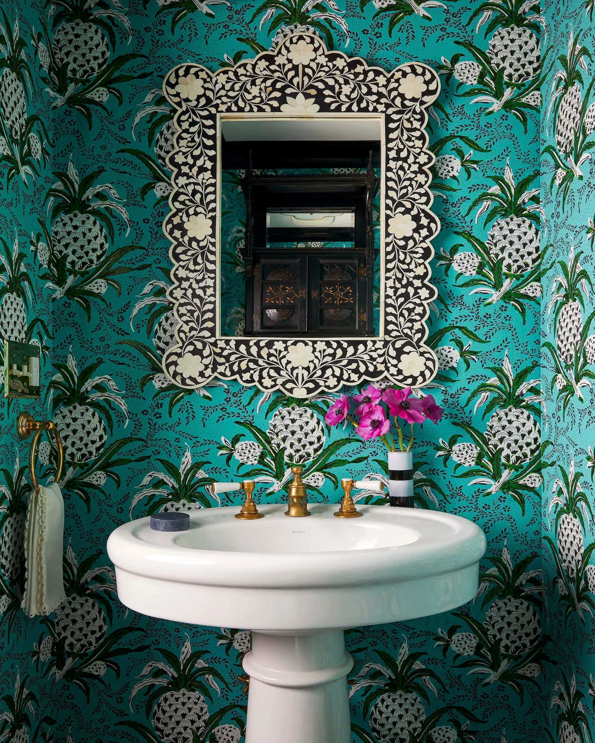 A white pedestal sink with brass fixtures sits against turquoise pineapple-print wallpaper under an ornate black and white inlay mirror.