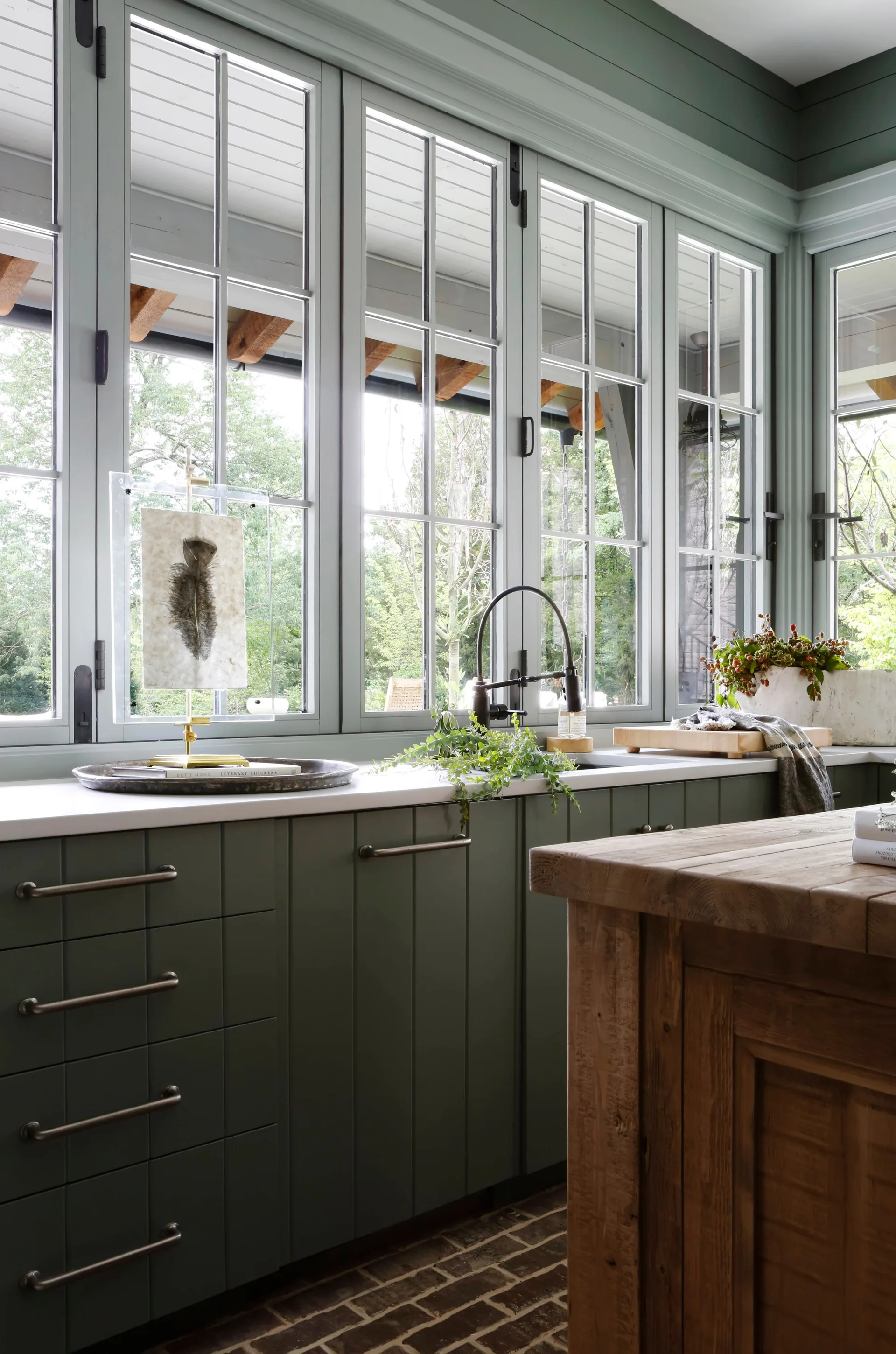 A green kitchen counter with a black faucet sits beneath tall, mullioned windows overlooking trees, next to a rustic wood island and brick floor.