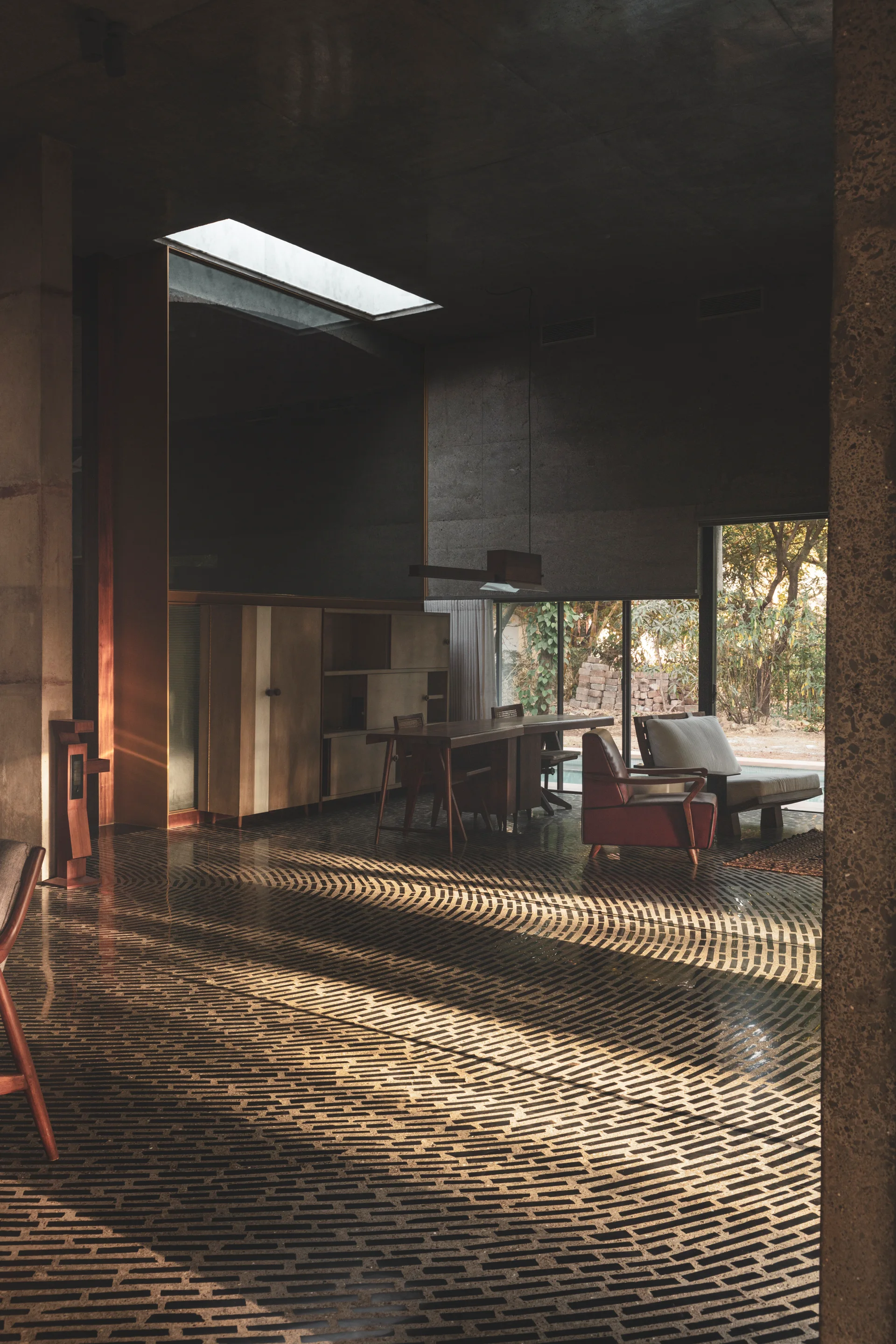 Mid-century modern wooden furniture sits on a patterned dark brick floor in a concrete room illuminated by a skylight and a large window.