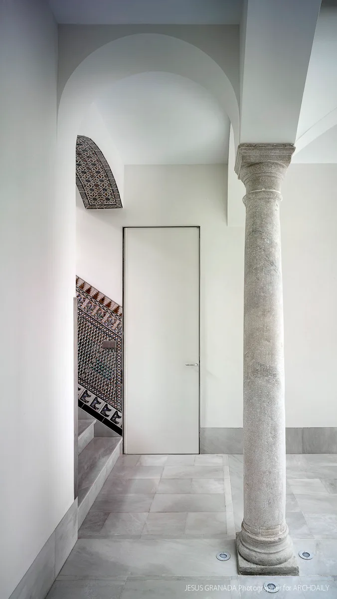 A minimalist entryway featuring a tall stone column, a simple white door, and a staircase with decorative blue and terracotta patterned tiles.