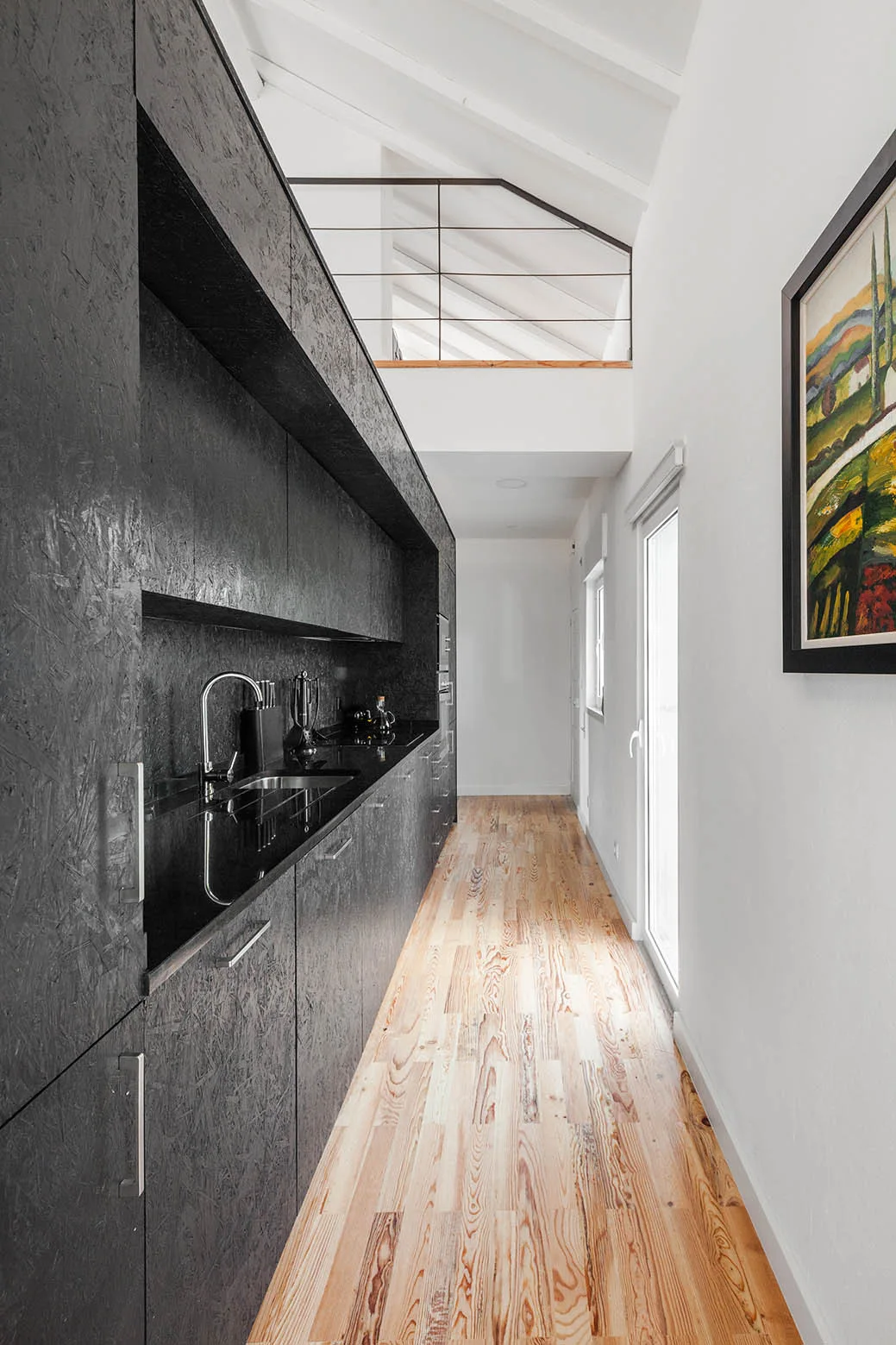 A narrow galley kitchen features textured black cabinets, a black countertop with a chrome faucet, and light wood plank floors under a high white ceiling.