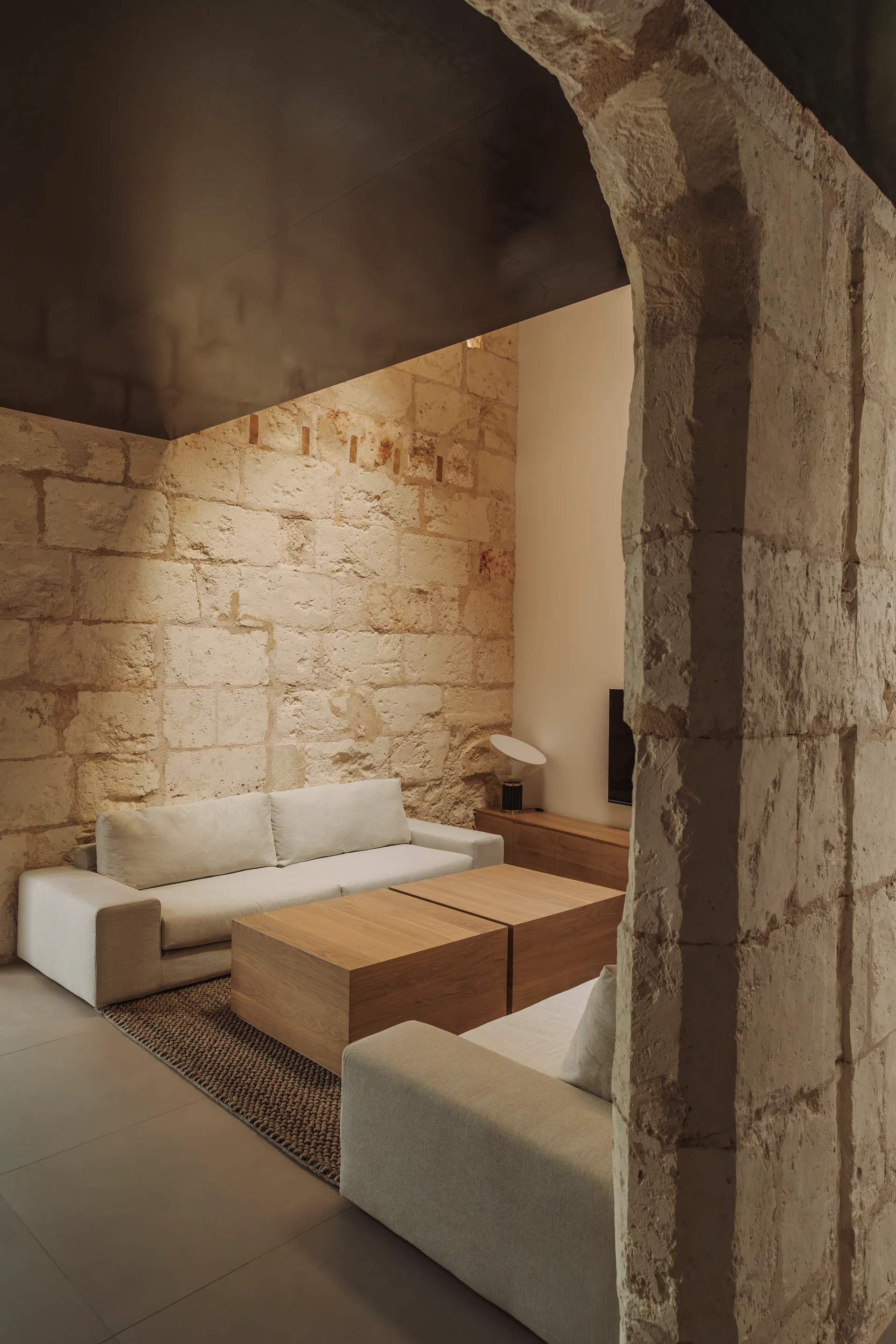 A minimalist living room seen through a stone archway, featuring an off-white sofa, two wooden block coffee tables, and a textured area rug.