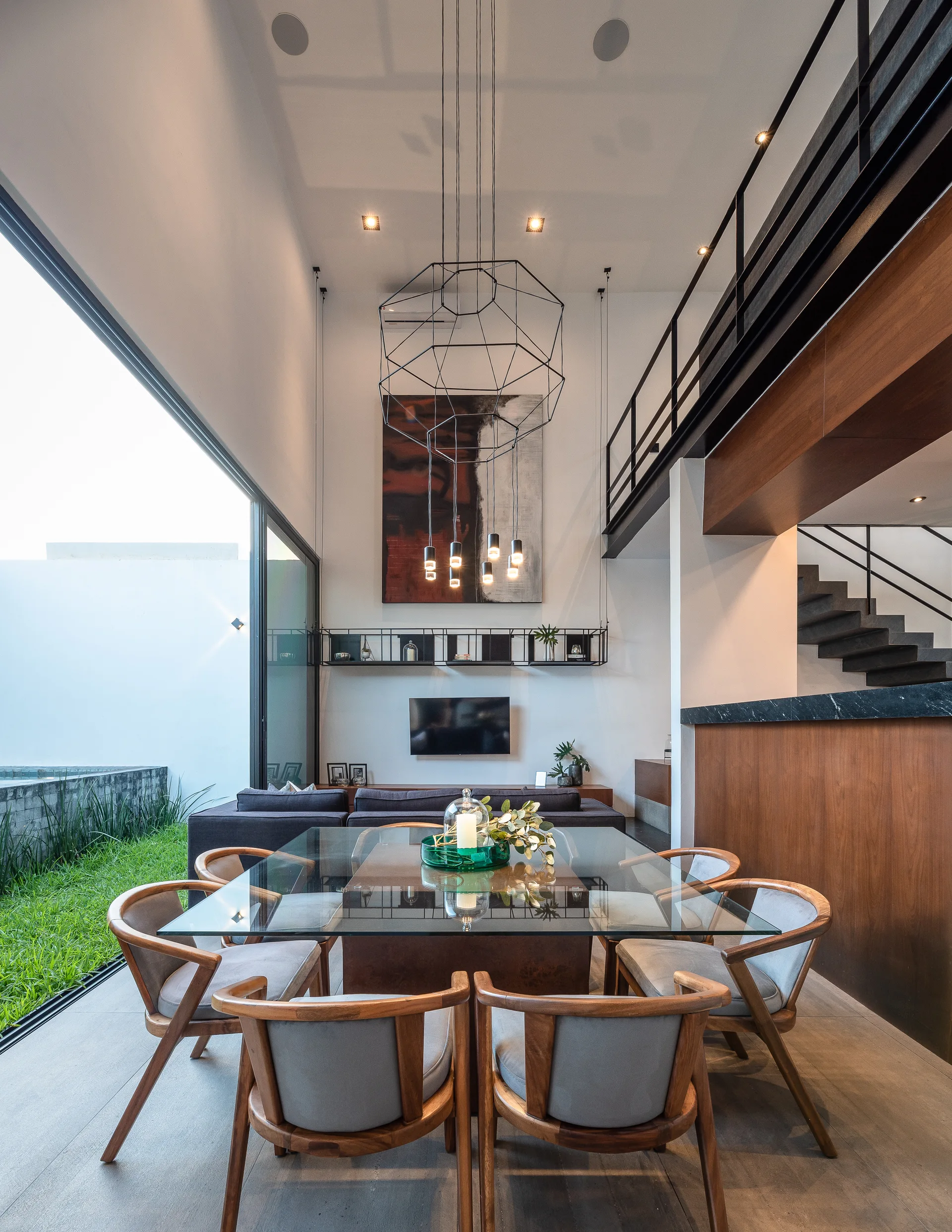 A modern dining room features a glass-topped table and curved wooden chairs beneath a large wire pendant light in a high-ceilinged open space.