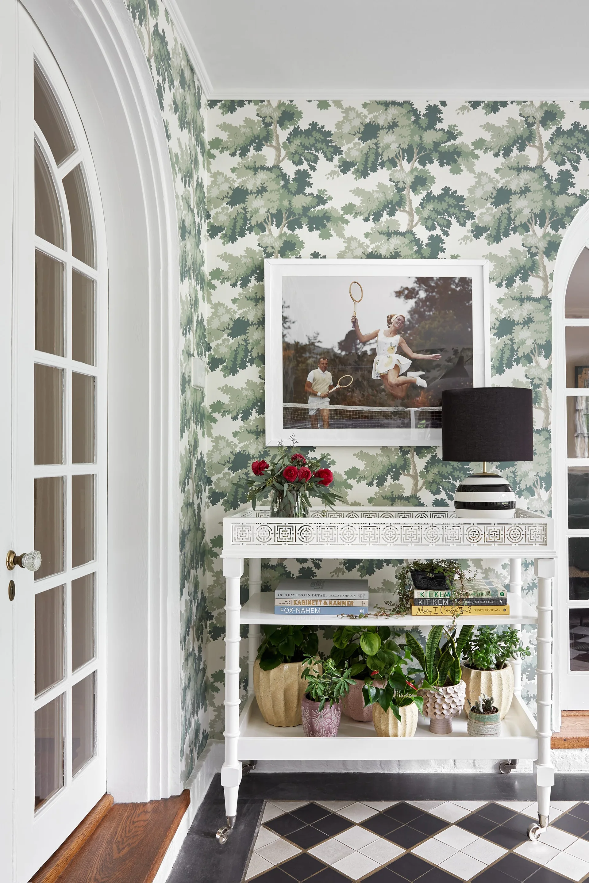 A white tiered console table with a black and white striped lamp sits against green tree wallpaper in a foyer with a checkerboard floor.