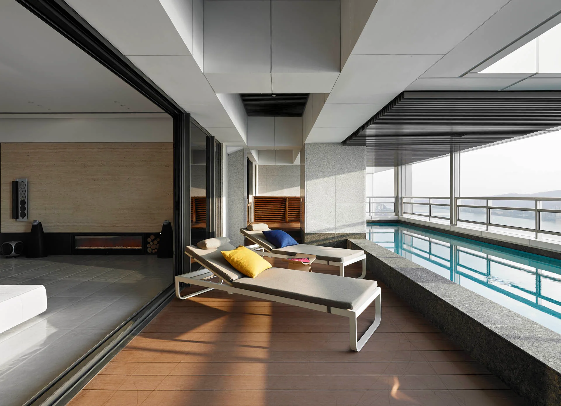 Modern apartment terrace with two lounge chairs on wood decking next to a long balcony lap pool and large sliding glass doors.