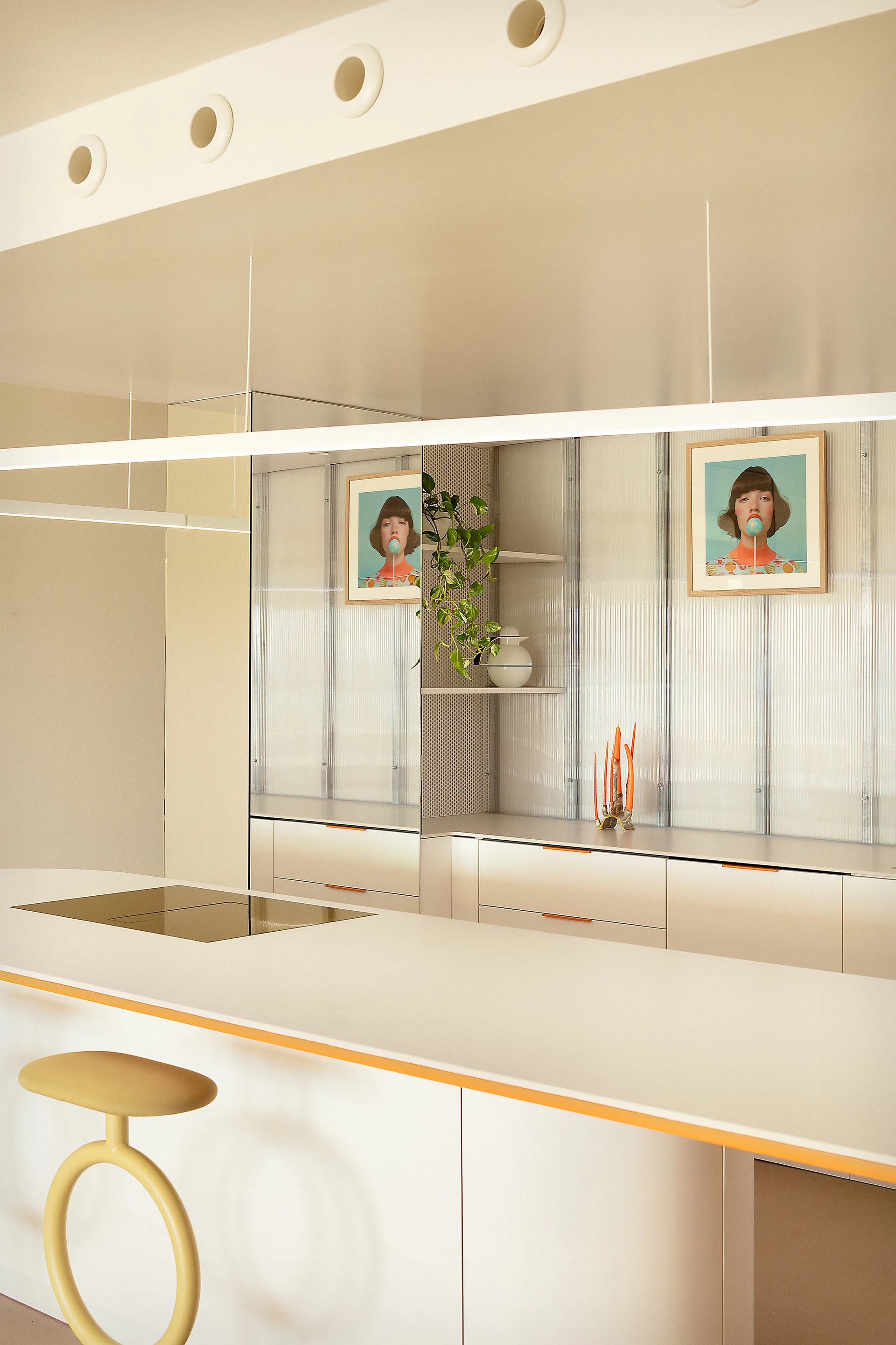 A modern cream kitchen featuring a white island with orange trim, a yellow sculptural stool, and a backlit fluted glass back wall.