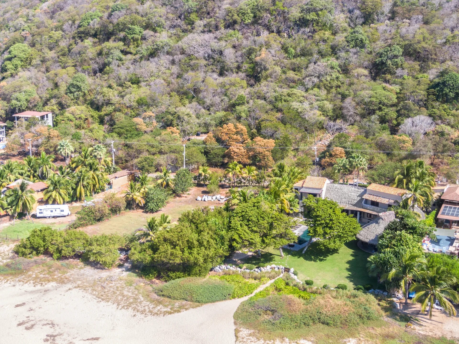 An aerial view of a beachfront home featuring a swimming pool, green lawn, and thatched roofs surrounded by palm trees and a forested hill.