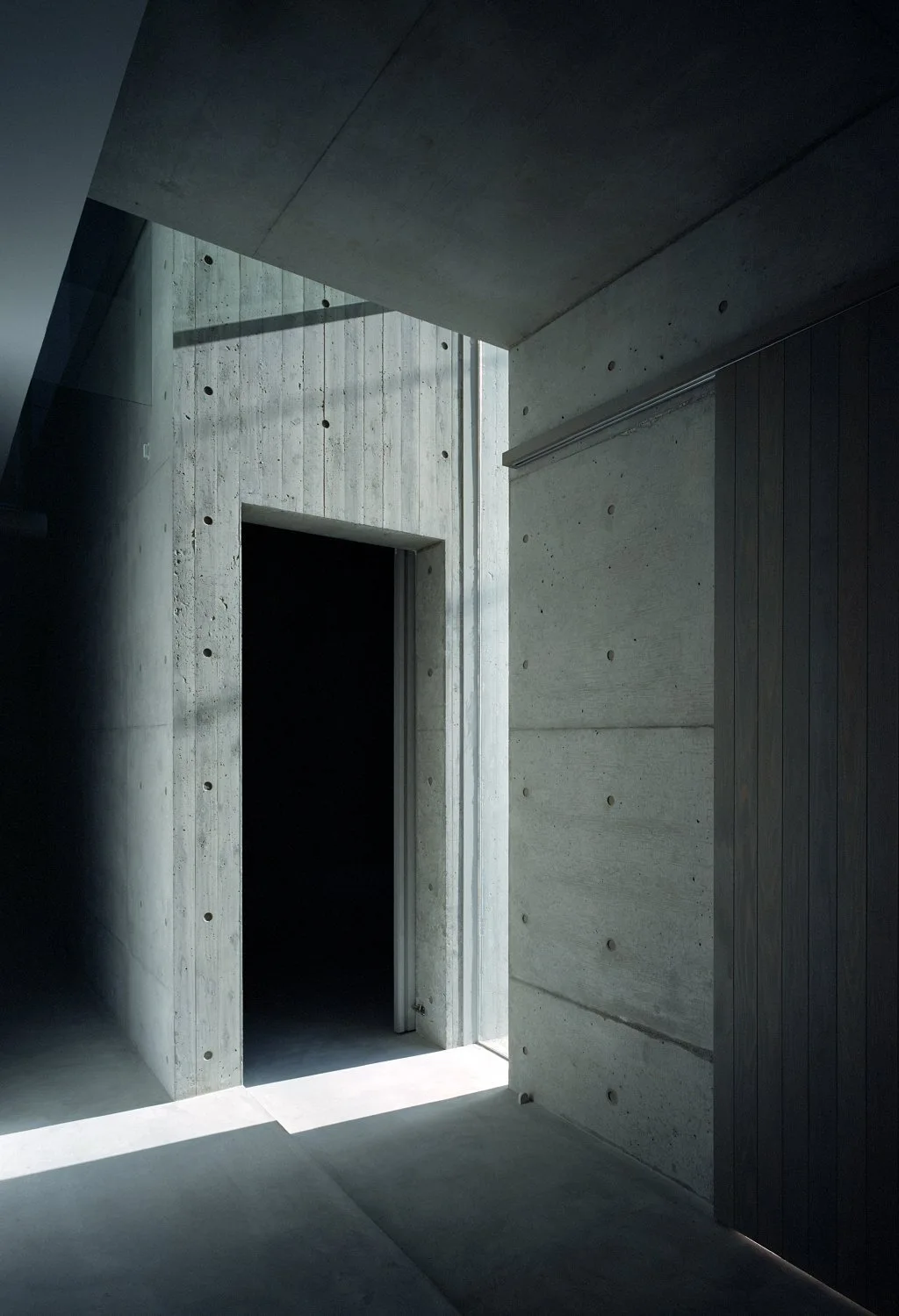 A minimalist hallway features textured board-formed concrete walls, a dark wooden sliding door, and sharp shadows from natural light.