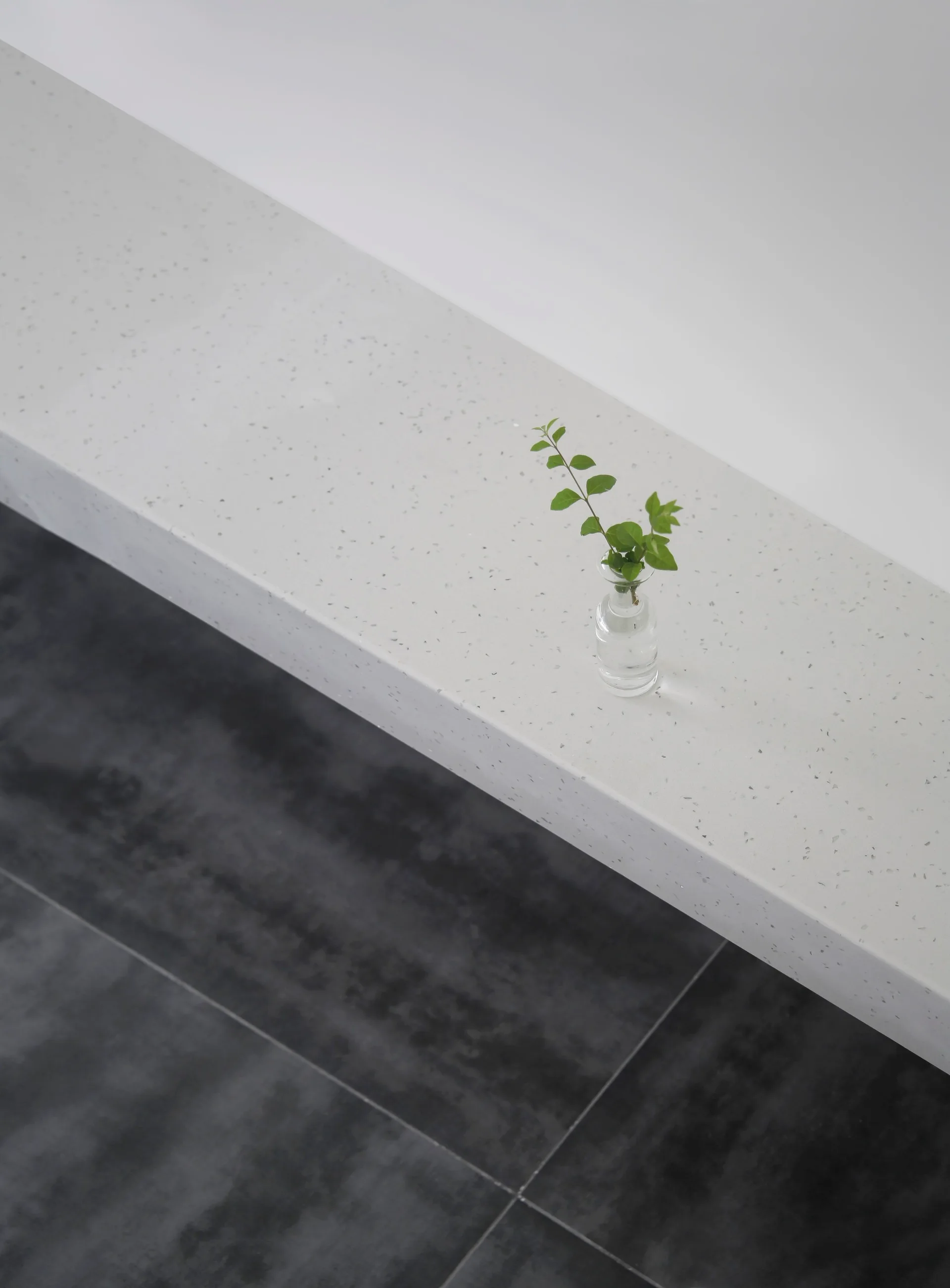 A high-angle view of a speckled white stone shelf holding a small glass vase with a green sprig above dark gray floor tiles.