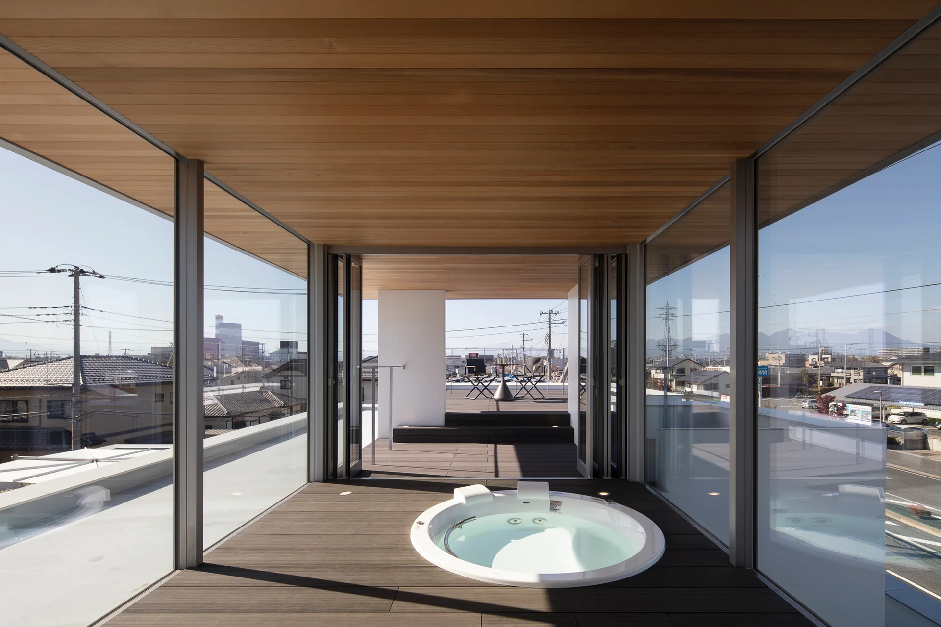 A modern glass-enclosed terrace features a circular white hot tub built flush into dark wood decking under a wood paneled ceiling.