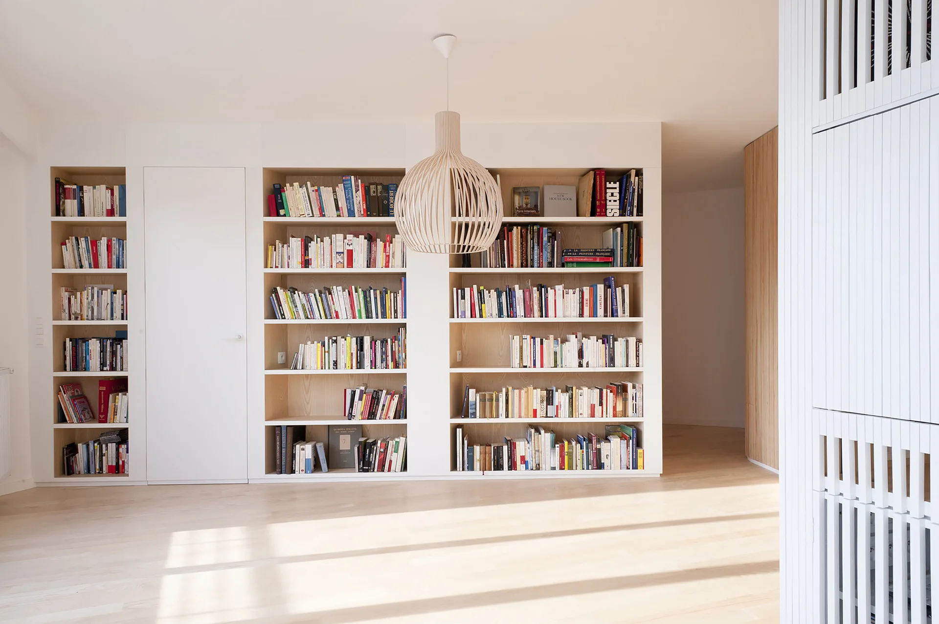 Floor-to-ceiling built-in wooden bookshelves line a bright room with light floors, a flush white door, and a spherical slatted pendant light.