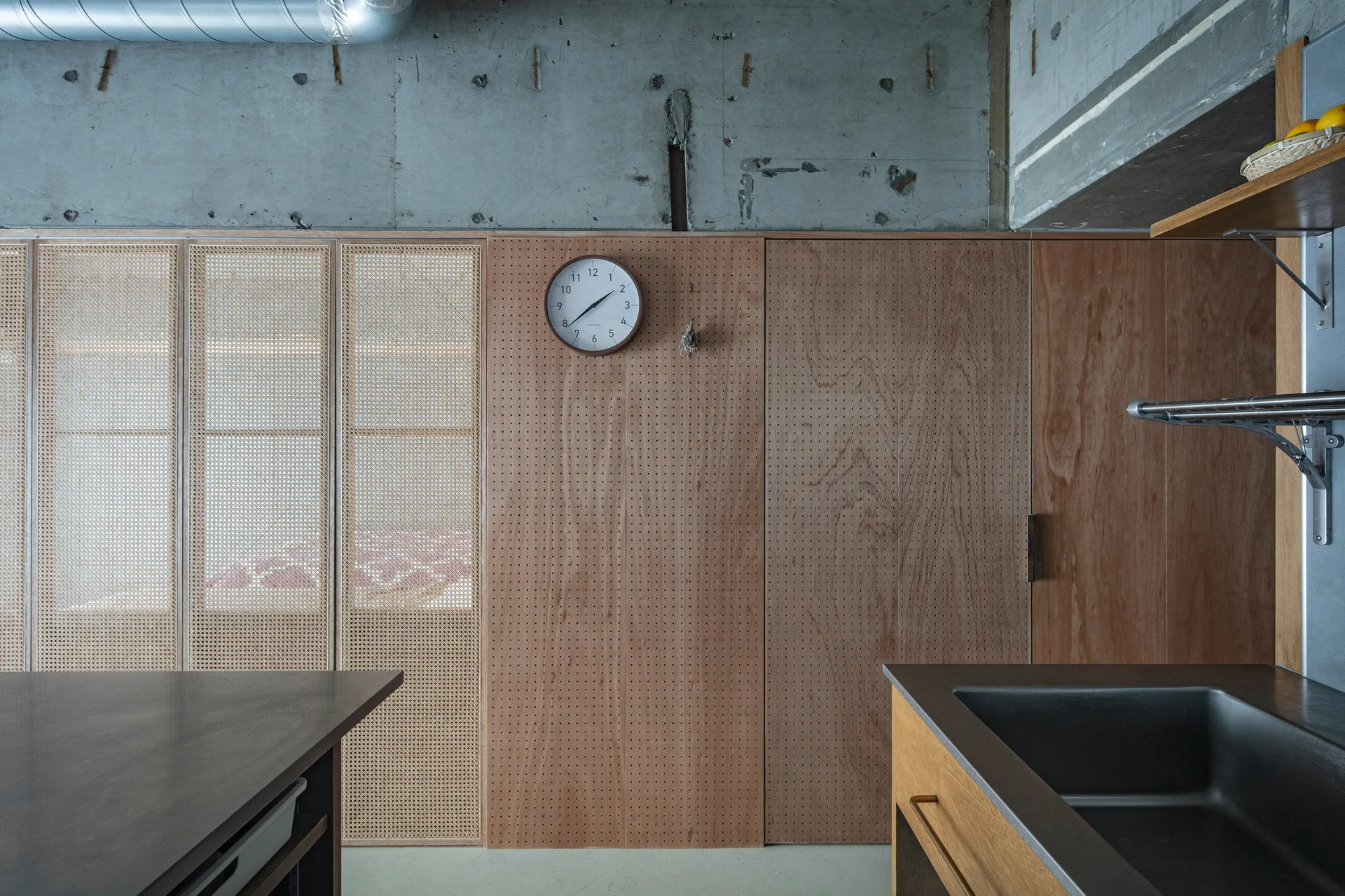 A kitchen featuring a raw concrete ceiling, a large wall of perforated wood panels, and sliding cabinet doors with woven rattan screens.