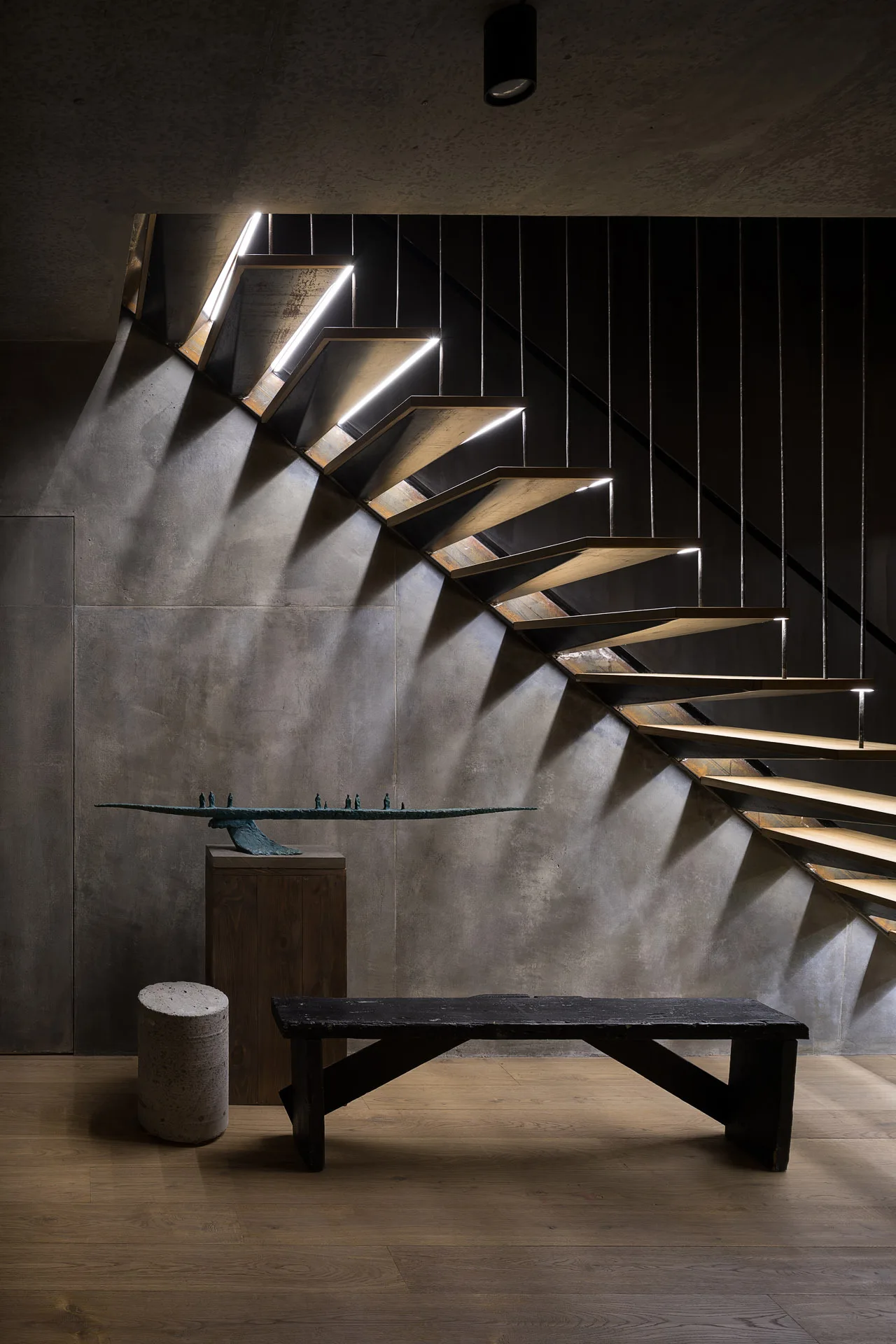 A floating metal staircase with integrated LED lighting under the treads stands against a raw concrete panel wall near a dark wooden bench.