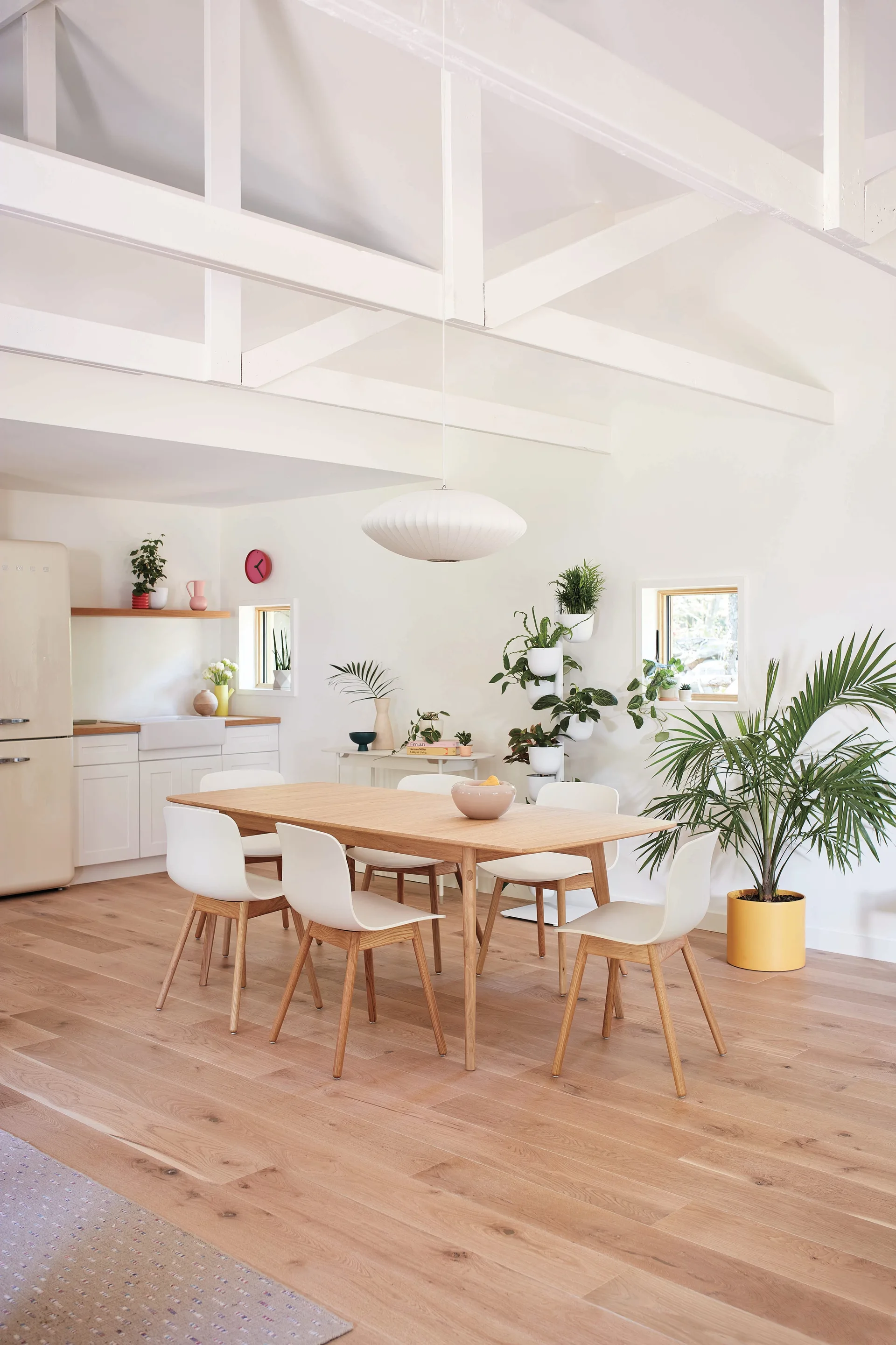 A bright dining room features a light wood table surrounded by white chairs under a high ceiling with exposed white beams and a saucer pendant light.