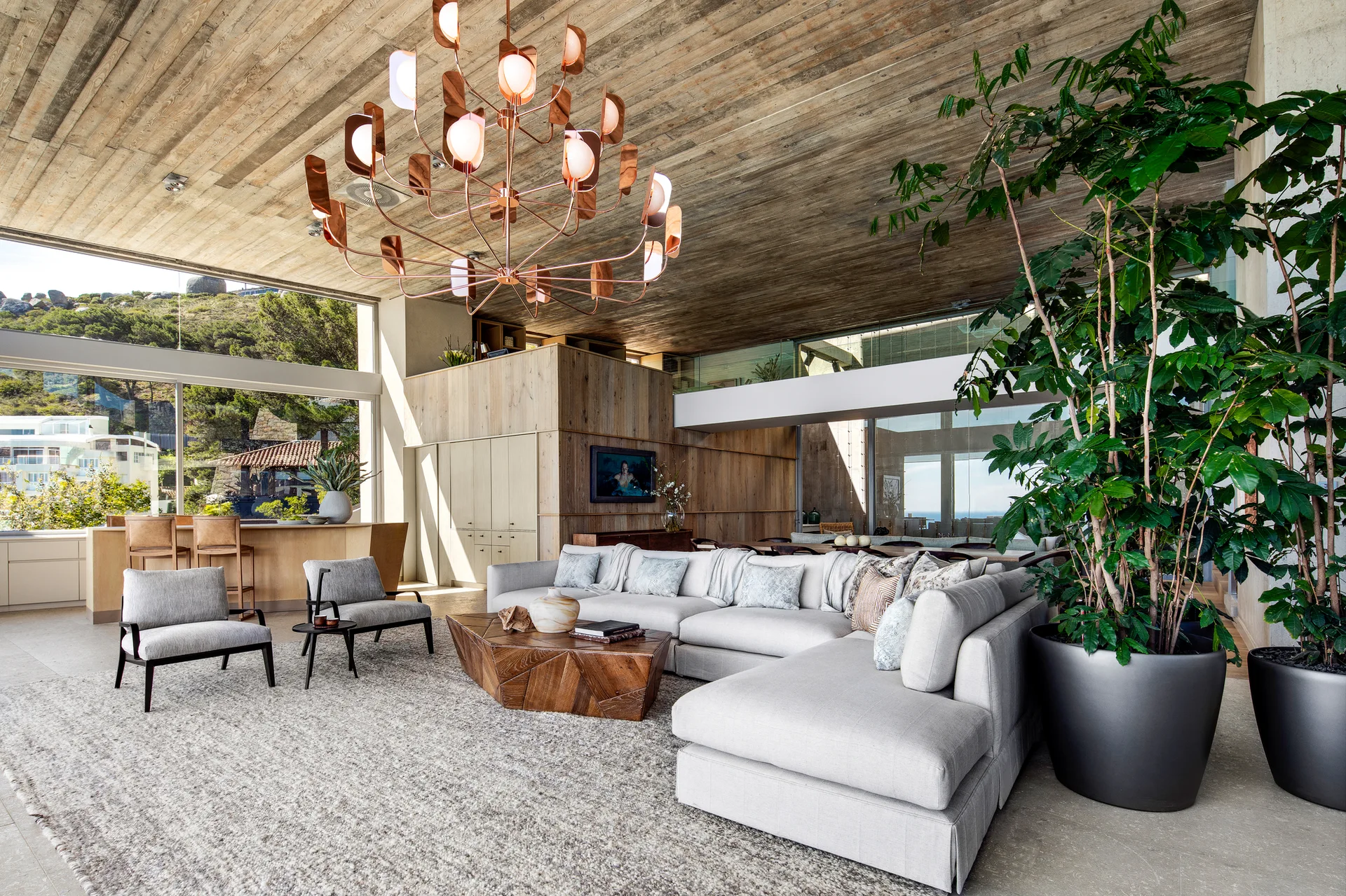A modern living room features a large gray sectional sofa, a faceted wood coffee table, a copper chandelier, and a wood plank ceiling.