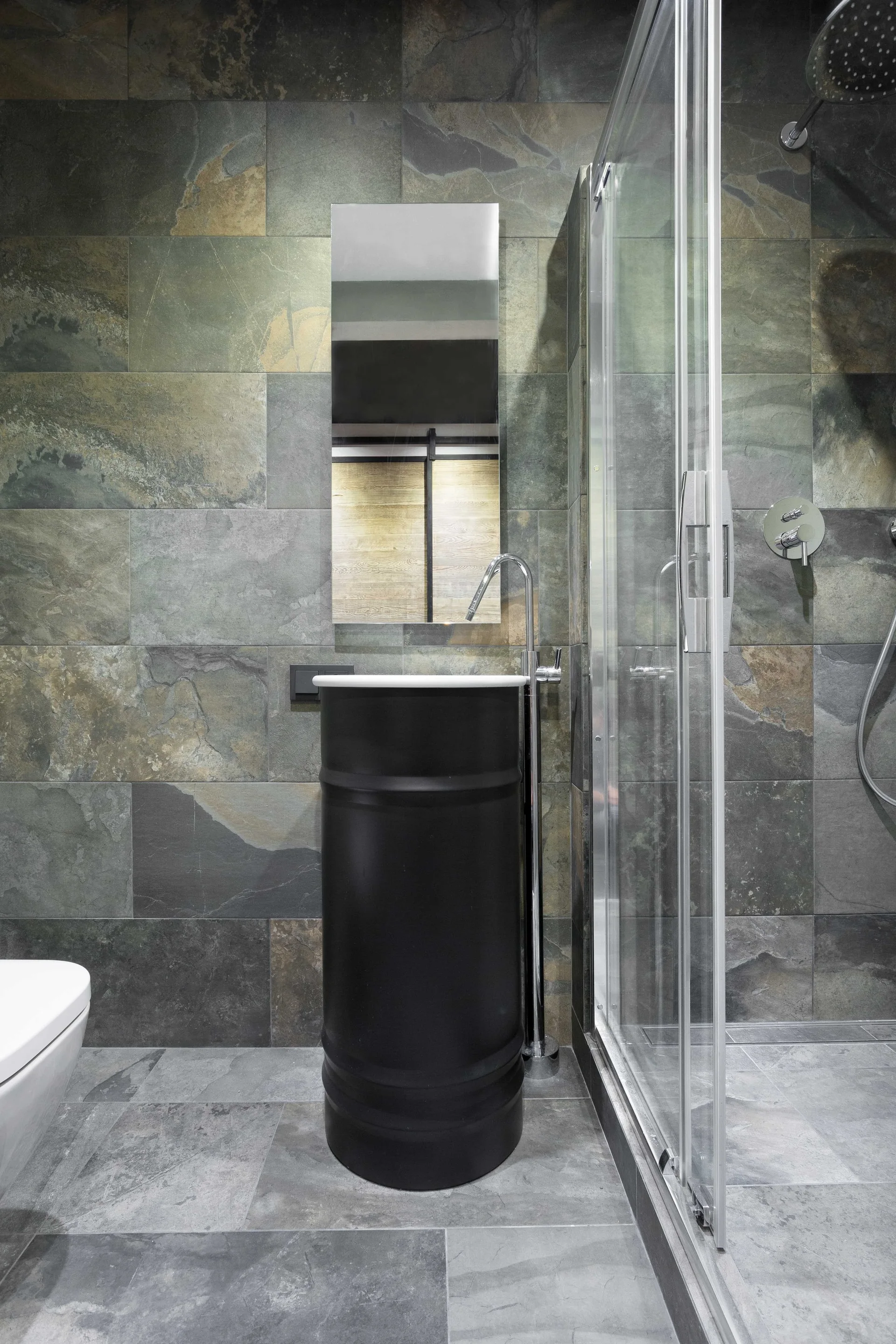 A modern bathroom features a black barrel-style pedestal sink with a tall chrome faucet against textured grey stone wall tiles.