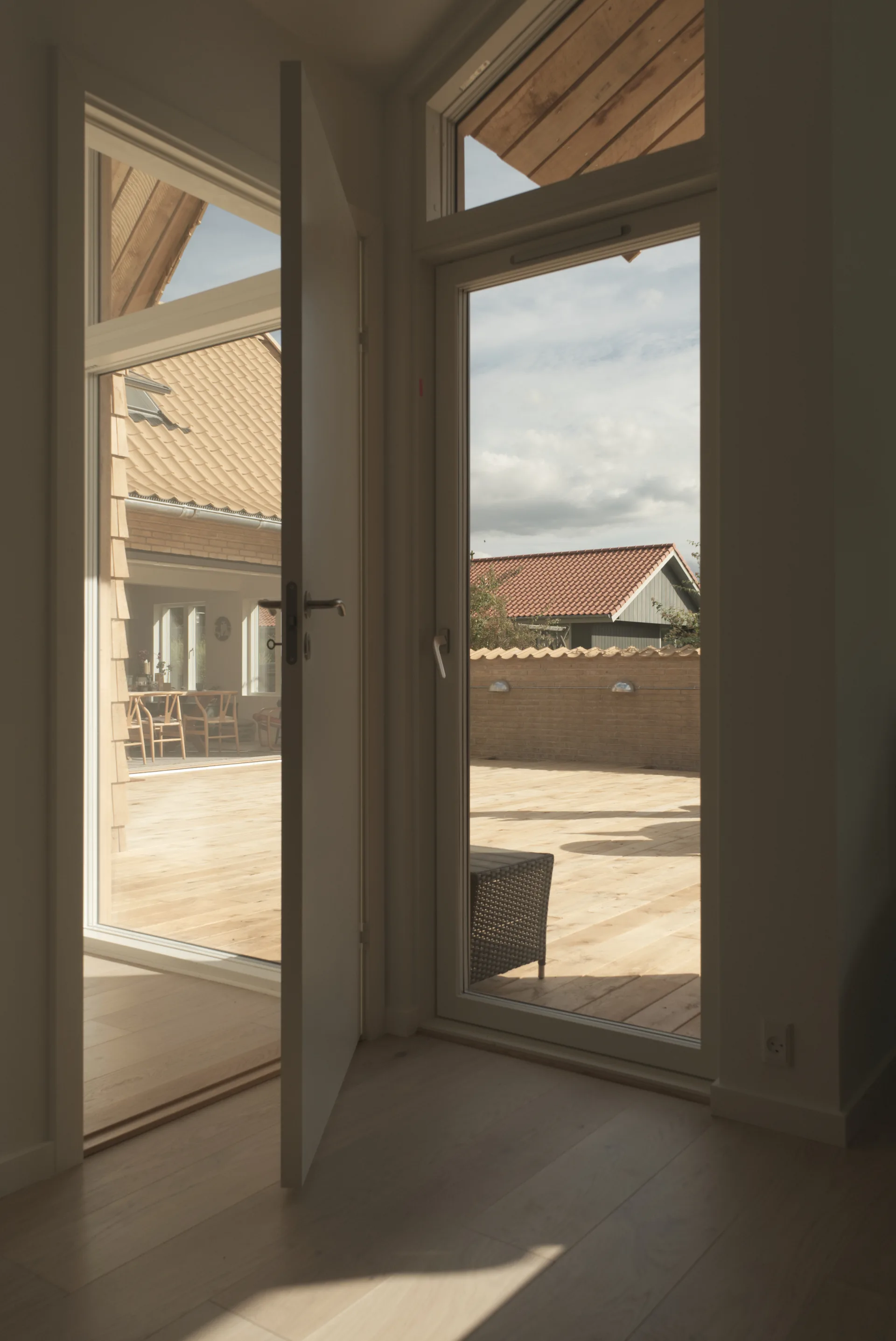 View from light wood indoor flooring looking through open white glass doors onto an outdoor wooden patio deck enclosed by a brick wall.