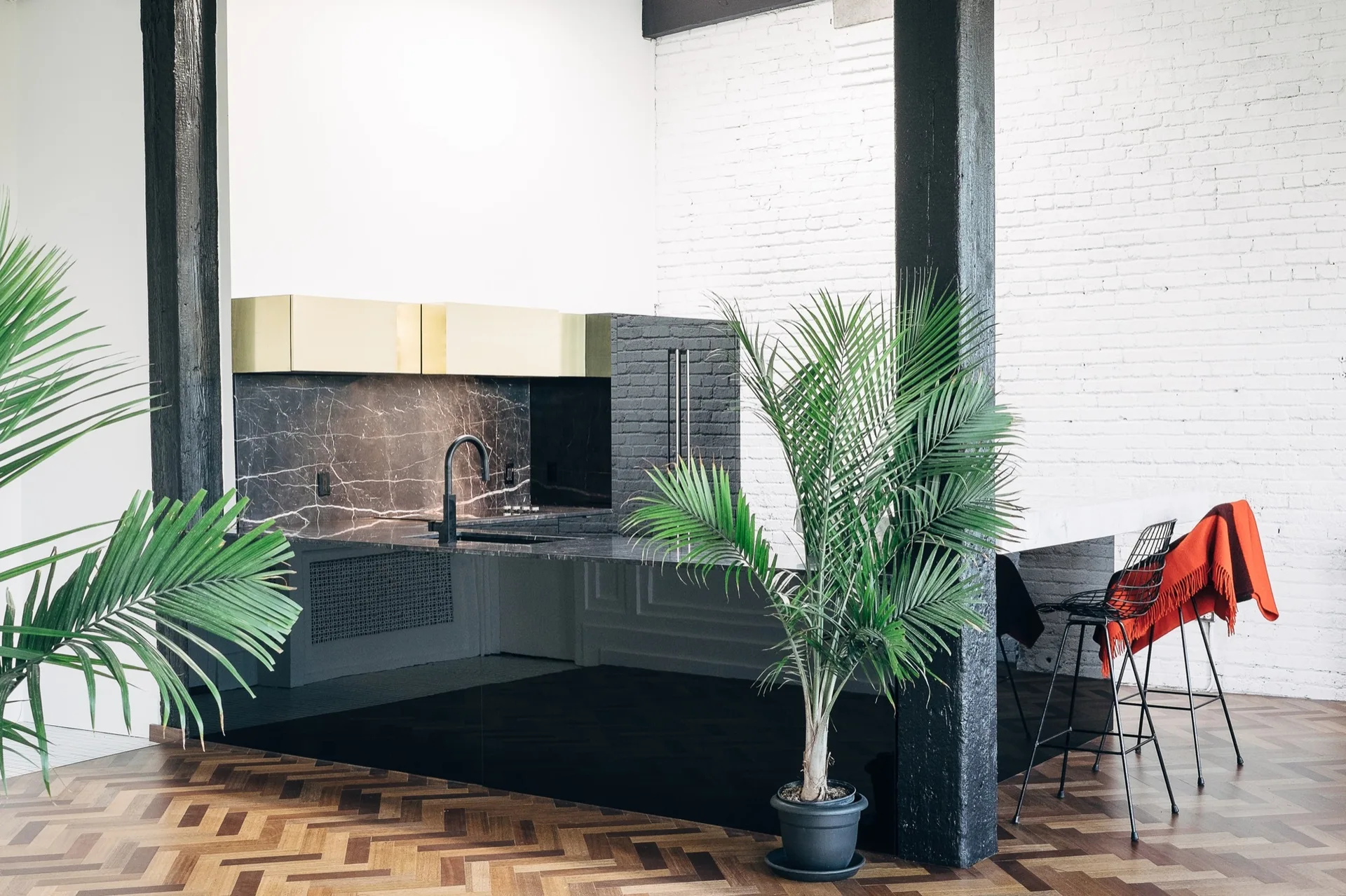 A loft kitchen featuring a black marble countertop, gold metal upper cabinets, painted white brick walls, and a black support column.