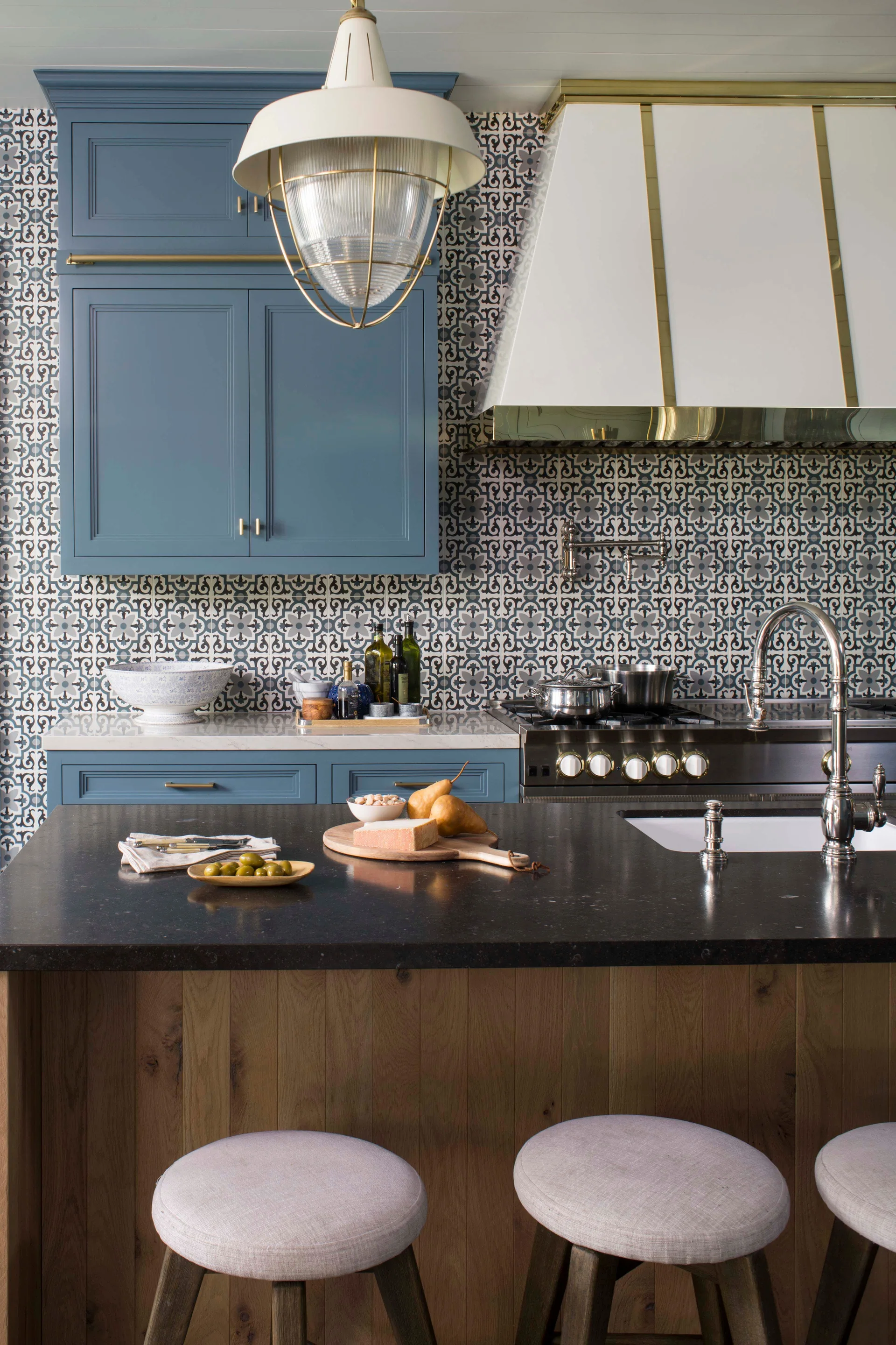 A kitchen features blue cabinets, a geometric patterned tile backsplash, a white and brass range hood, and a dark stone island countertop with bar stools.