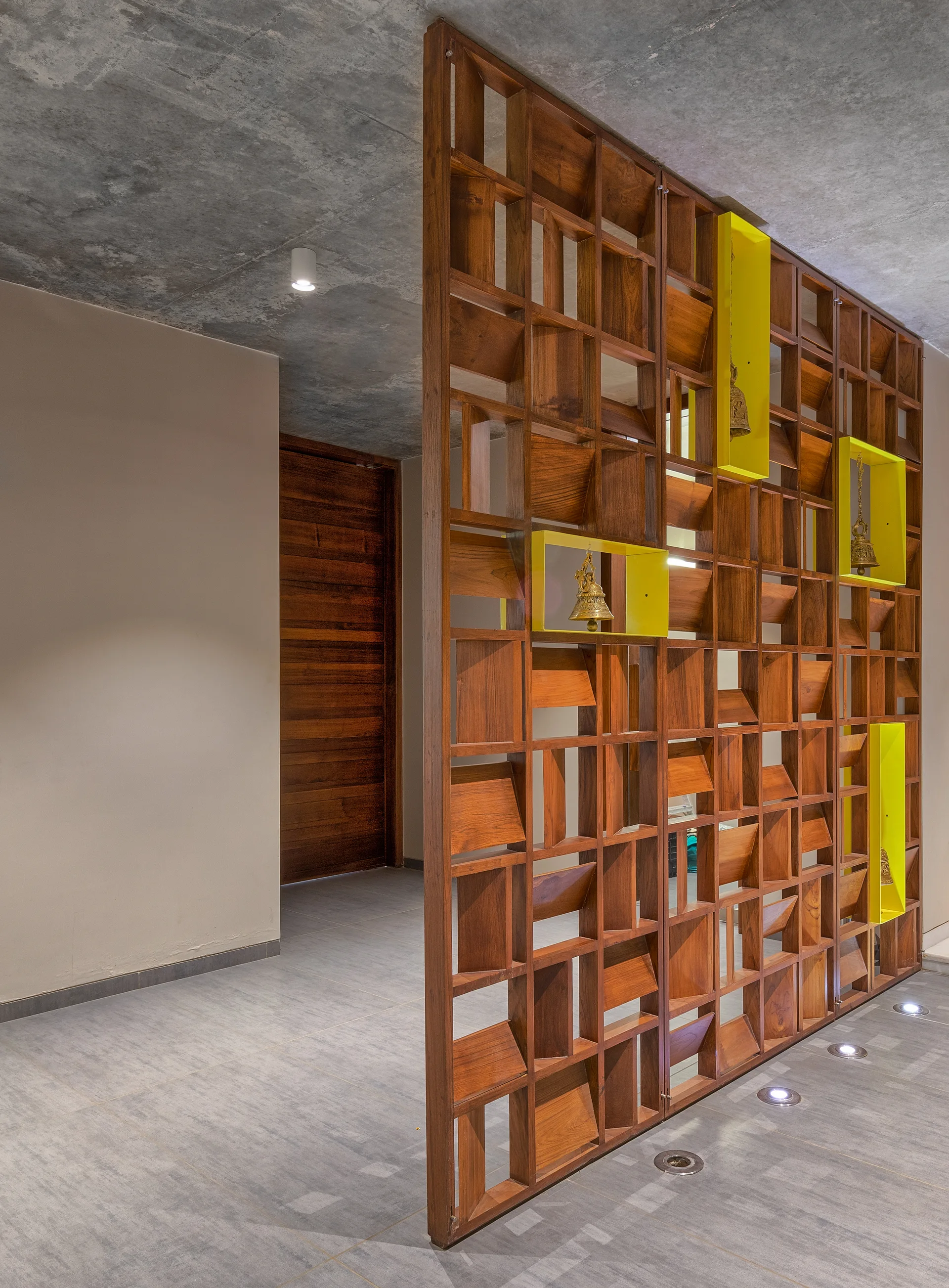 A tall wooden lattice room divider with yellow box displays stands on a grey tiled floor beneath an exposed concrete ceiling.