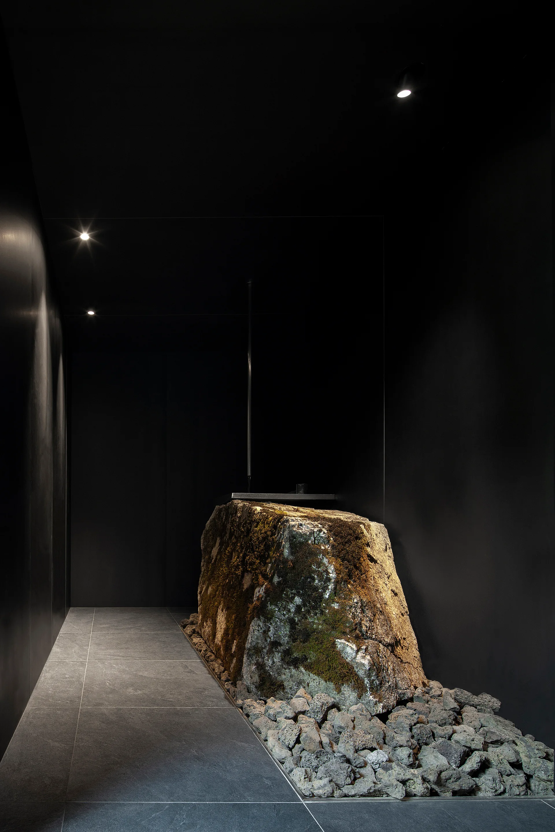 A minimalist dark hallway features a large moss-covered boulder surrounded by small grey stones under pinpoint ceiling spotlights.