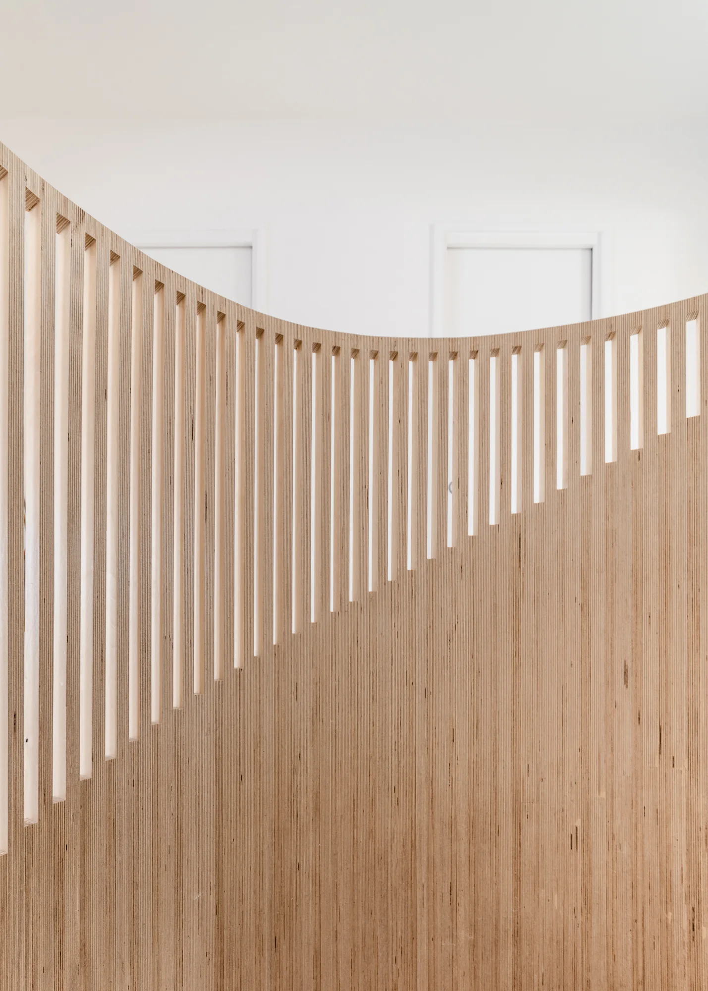 A close-up of a curved staircase balustrade featuring vertical light wood slats made of plywood against a plain white wall.