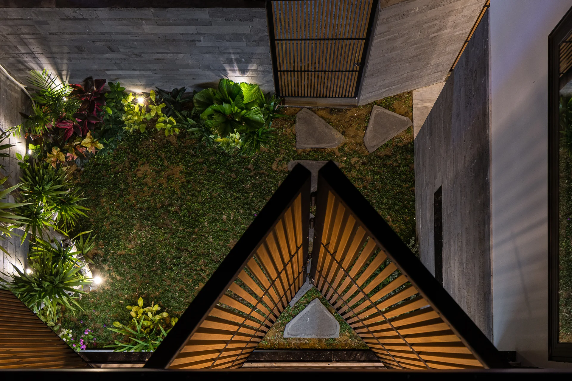 An overhead view of a courtyard garden with triangular stepping stones, lush border plants under spotlights, and slatted wooden screens.