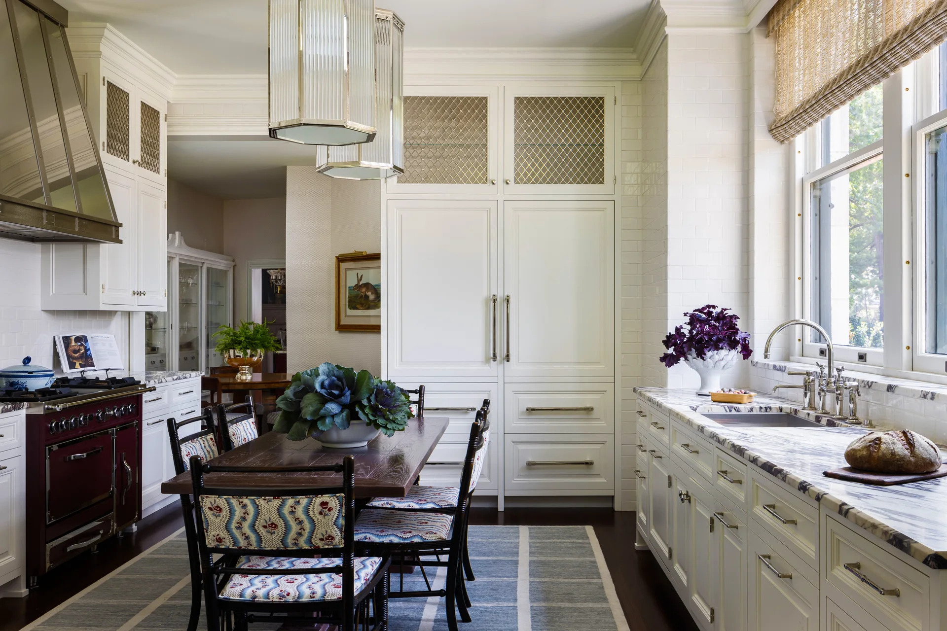 A traditional white kitchen features a central wooden dining table with patterned chairs, a burgundy range, and marble countertops.