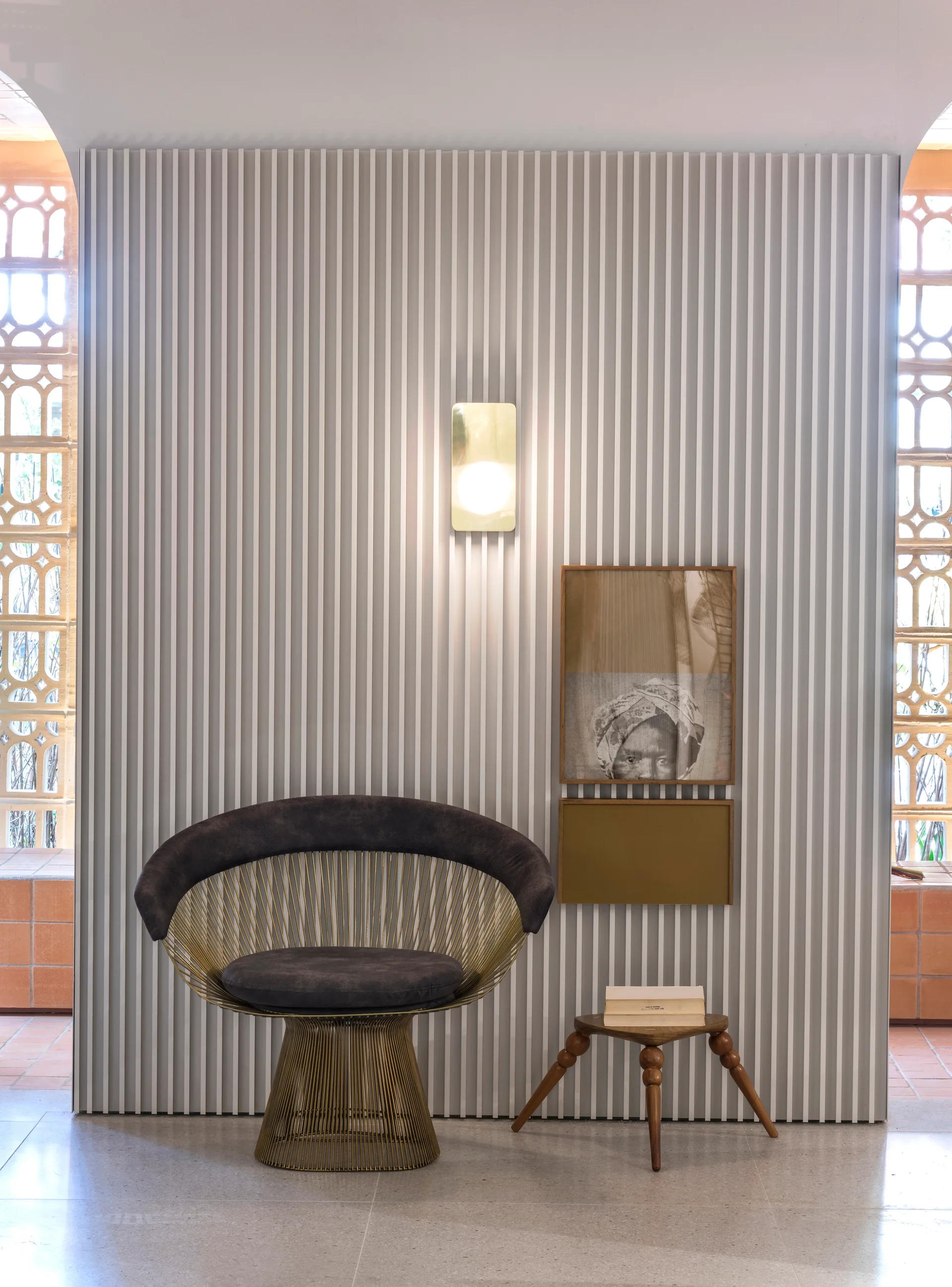 A circular wire-frame accent chair sits against a white vertical slat wall beside a small wooden stool, a warm wall sconce, and framed artwork.