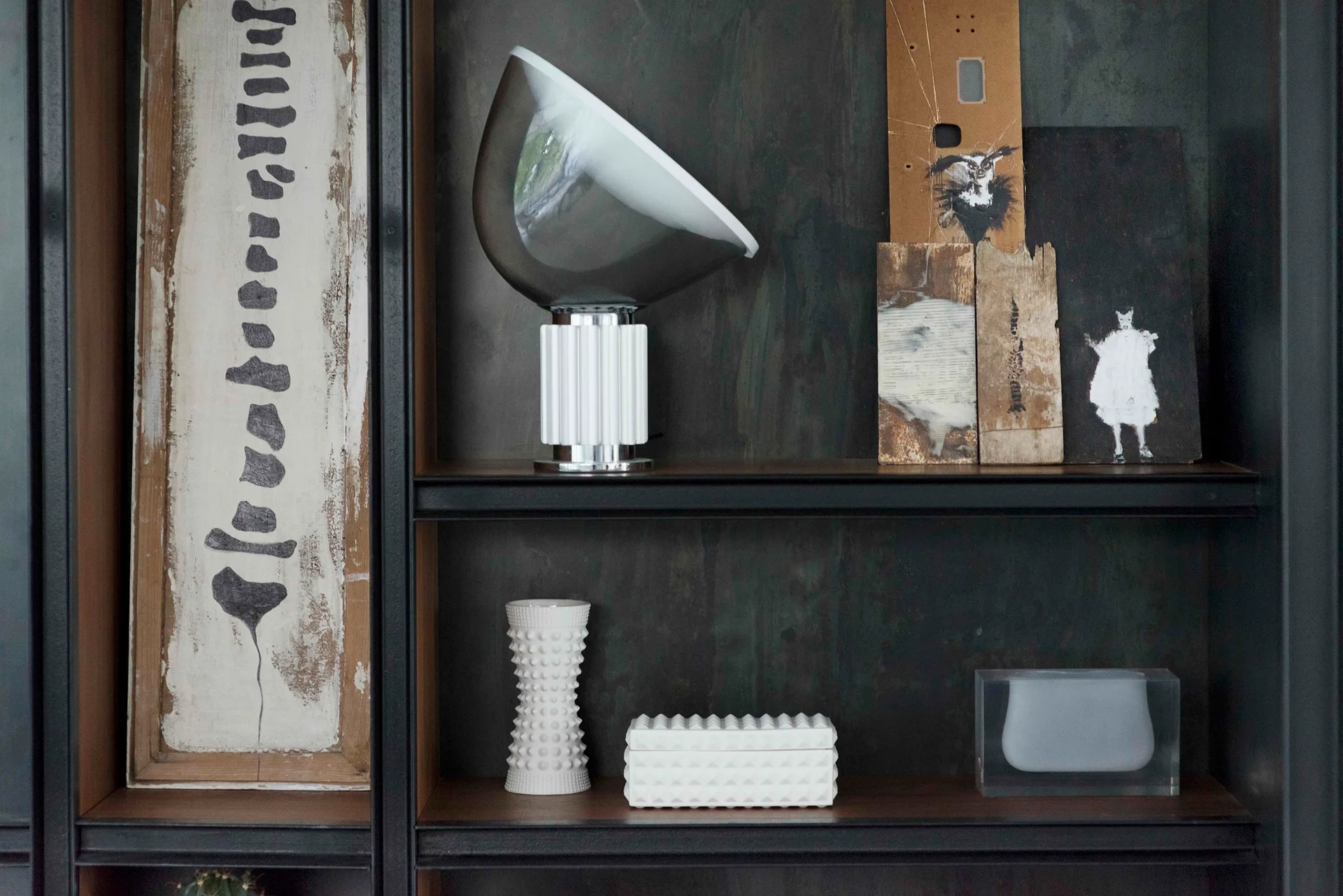 A dark metal and wood shelving unit displaying a modern table lamp, textured white ceramic objects, and small abstract paintings leaning against the back.