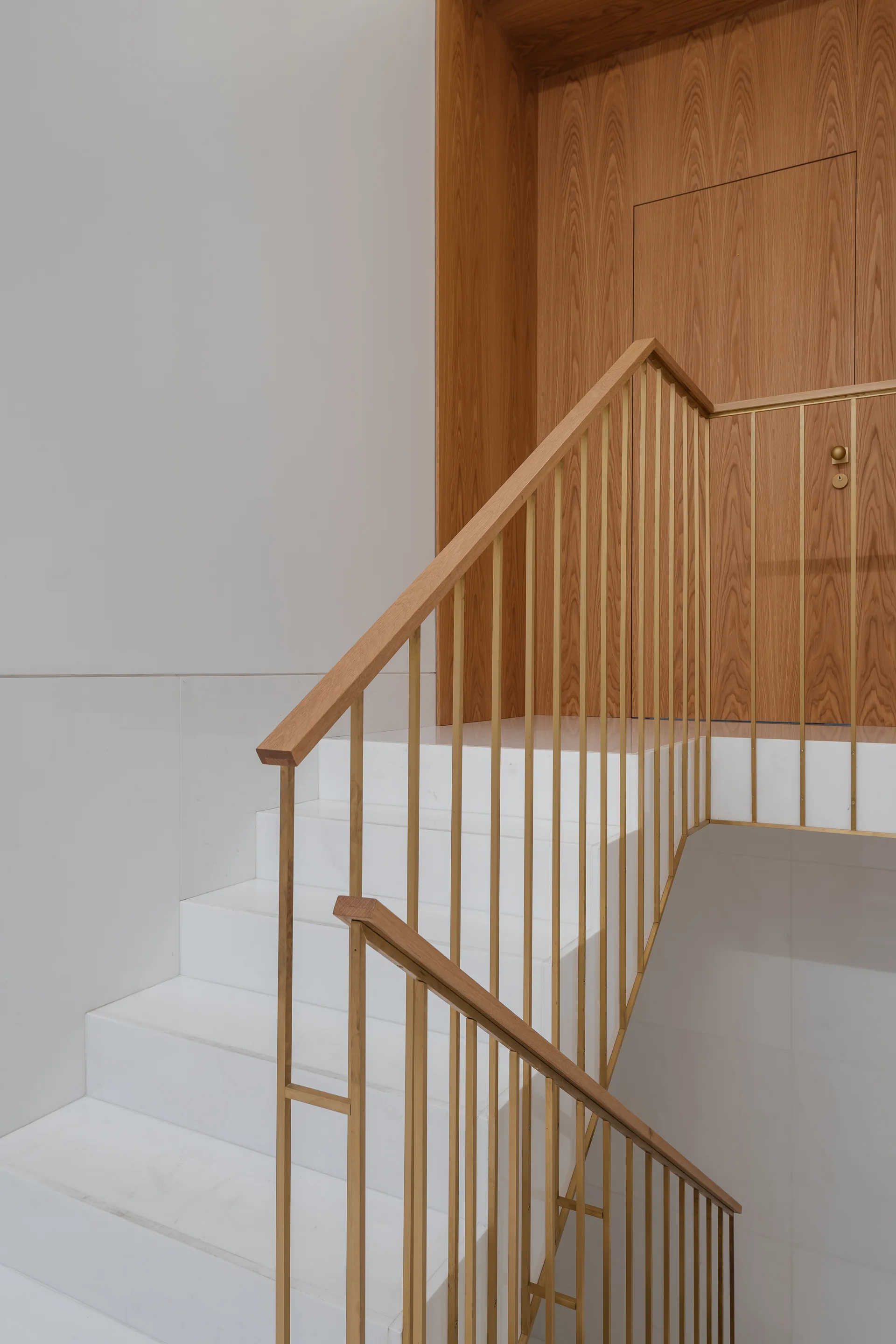 A modern white staircase features slim gold-toned metal balusters and a wooden handrail leading up to a flush wood-paneled wall and door.