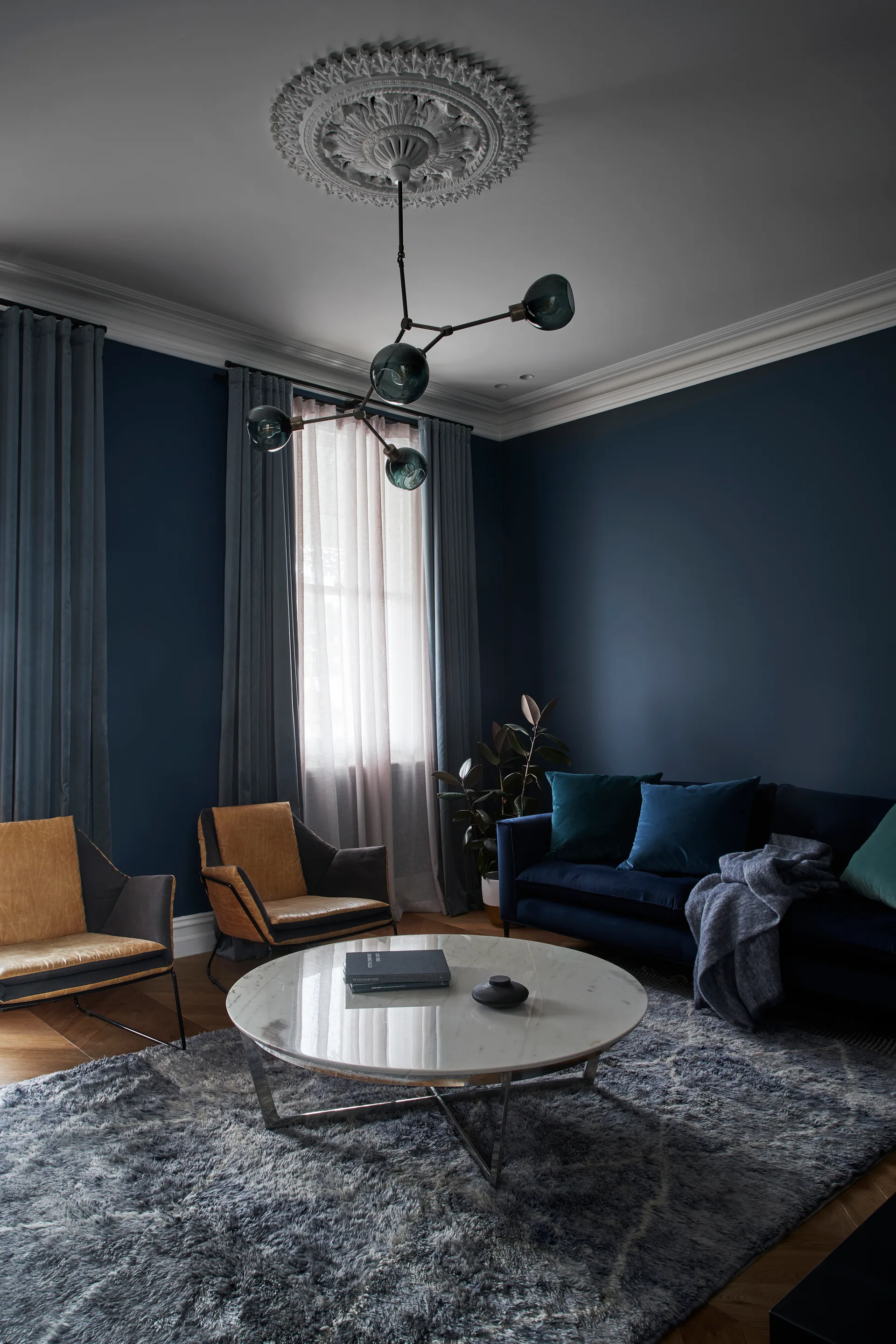 A living room featuring dark blue walls, a round marble coffee table on a plush grey rug, and a velvet sofa beneath an ornate ceiling medallion.