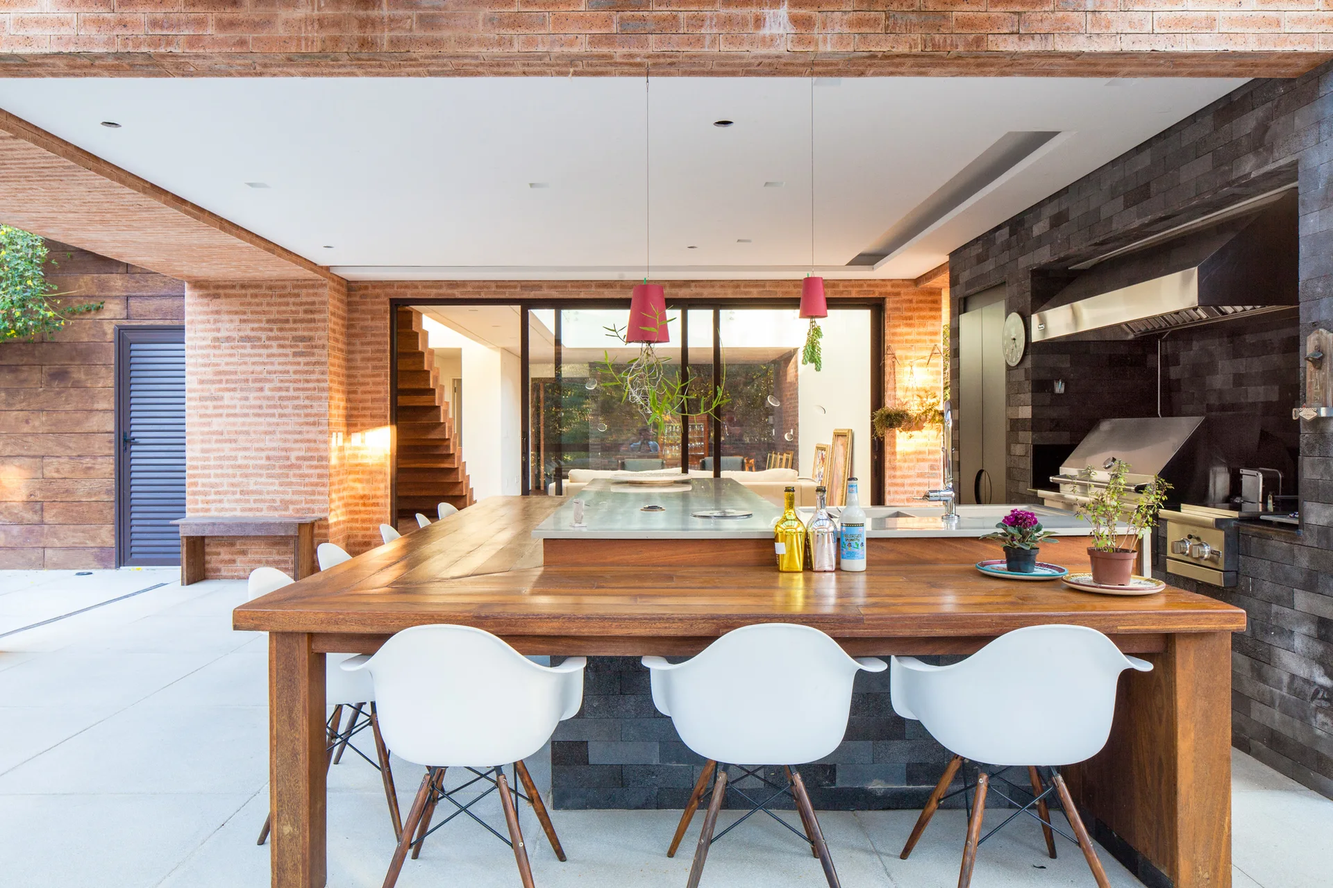 A covered patio features an outdoor kitchen with a dark stone grilling station, a long wooden dining table, white chairs, and red brick walls.