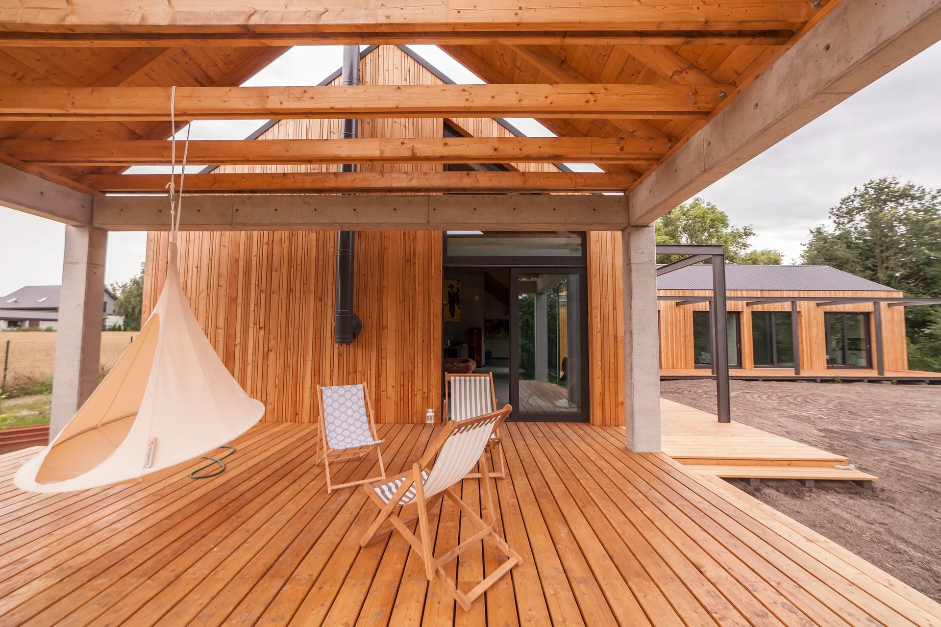 A covered wooden deck features a suspended canvas pod chair, two striped wood lounge chairs, and an overhead exposed beam structure.