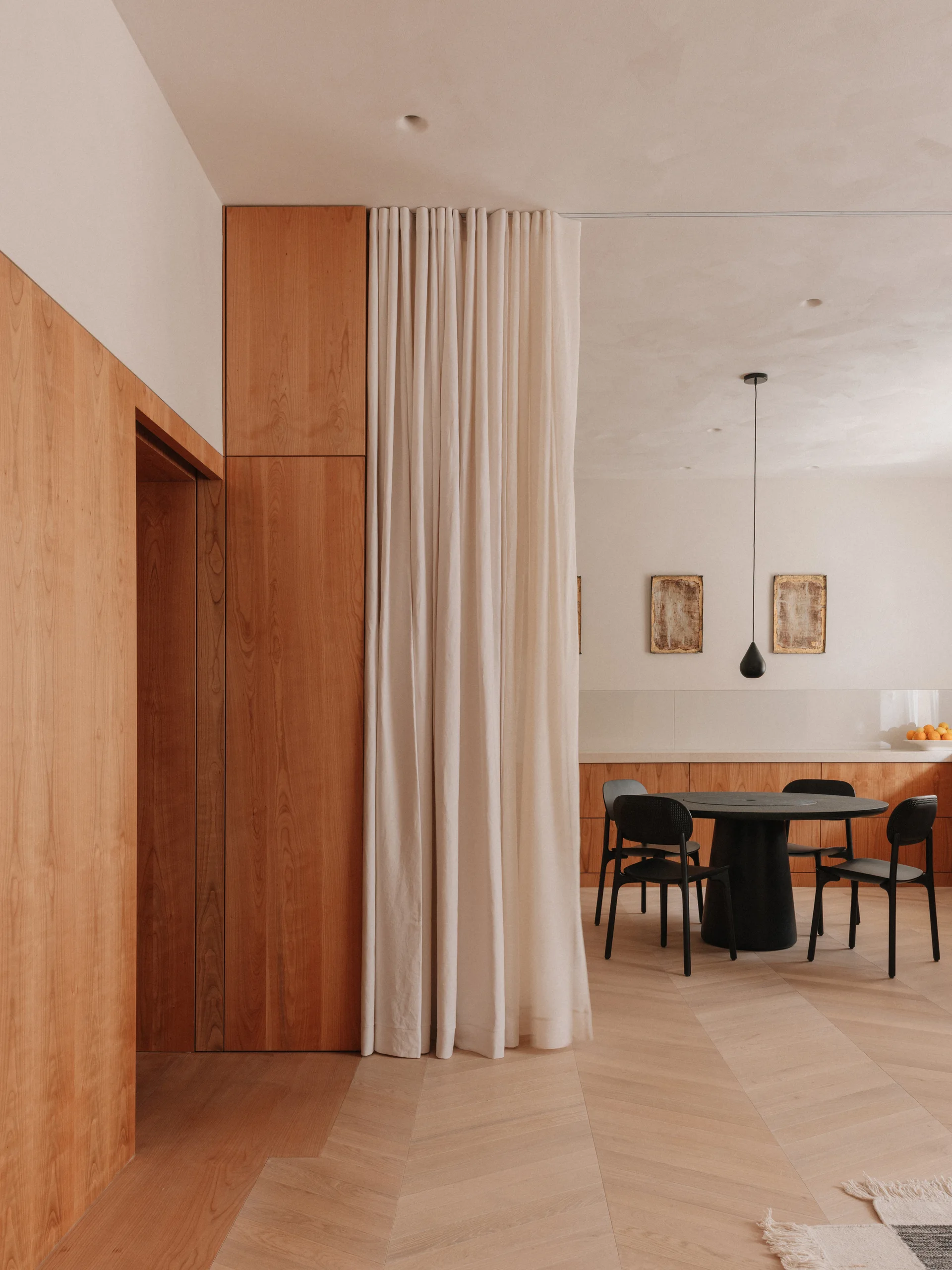 A minimalist dining room features a black round table, chevron wood floors, warm wood paneling, and a tall cream fabric divider curtain.
