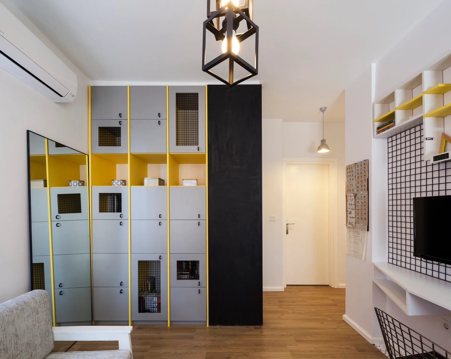 A modern home office features grey and yellow locker-style cabinets, a black wire wall grid above a desk, and a geometric pendant light.