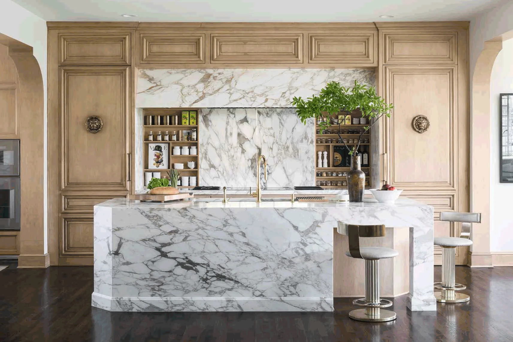 A kitchen with light wood paneled cabinetry features a large veined marble island, matching marble backsplash, and two upholstered swivel stools.