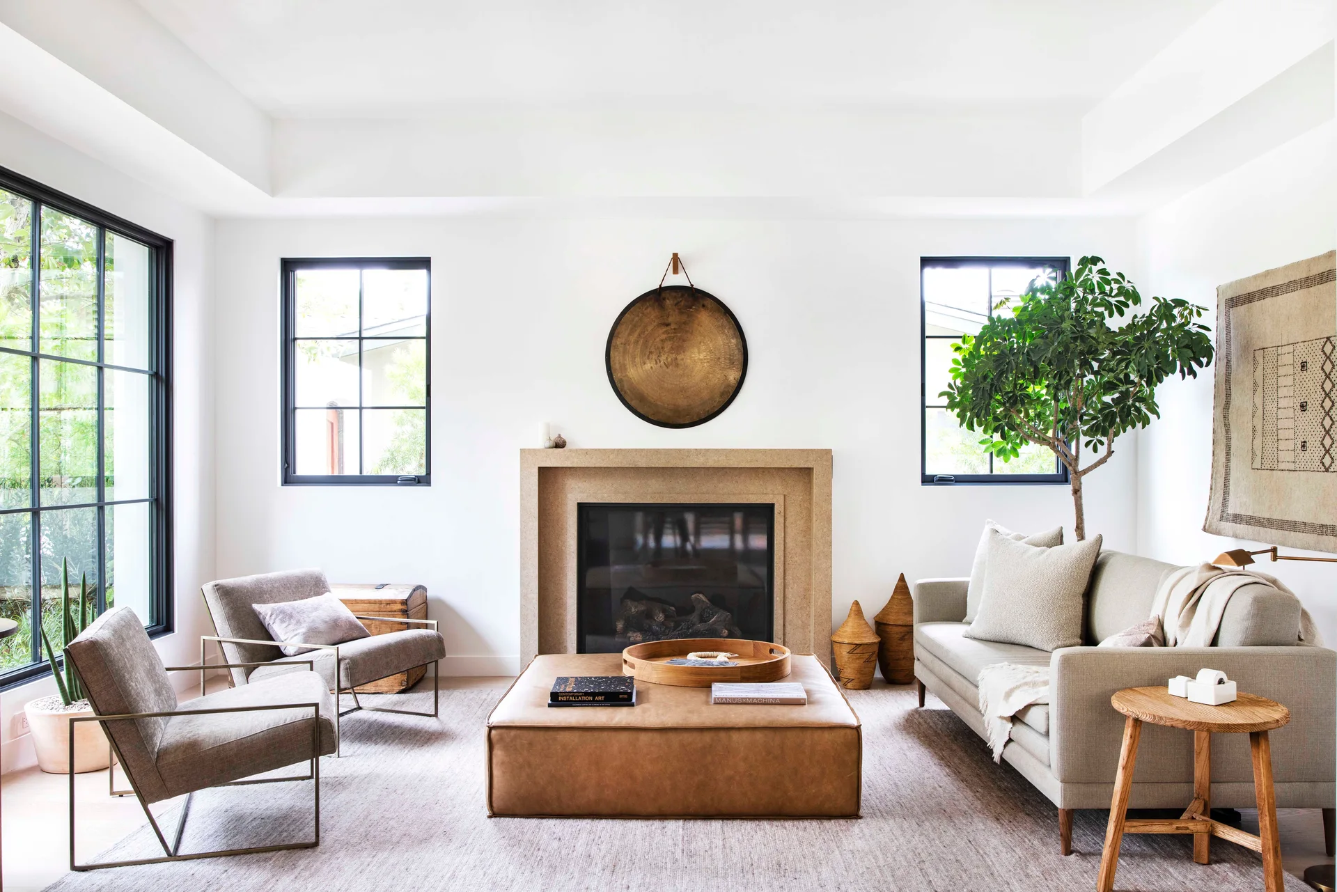 A bright living room with a stone fireplace flanked by black-framed windows, a large leather ottoman, and neutral upholstered sofas.