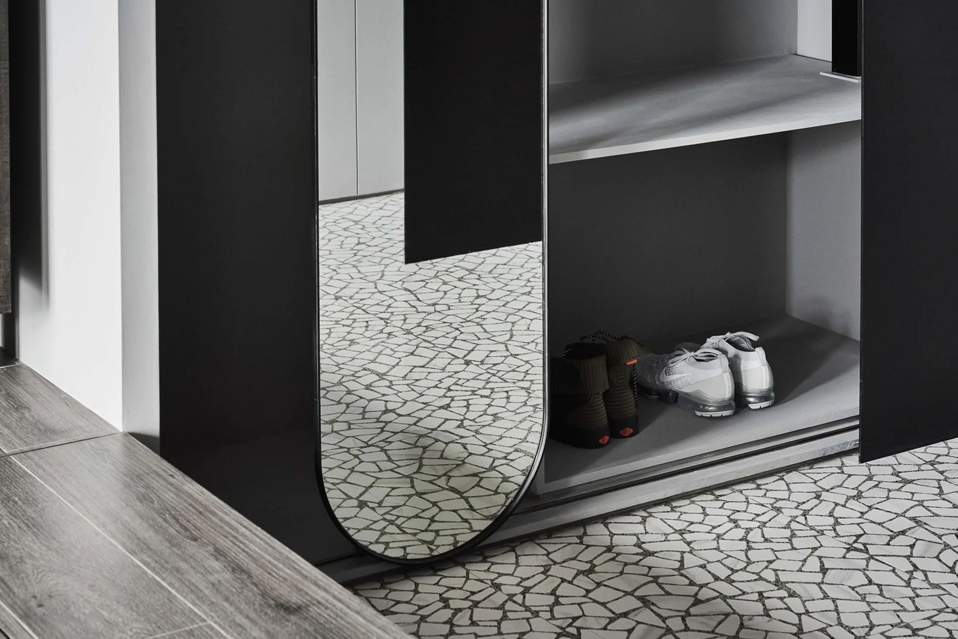 A minimalist closet unit with black framing, a tall oval-shaped mirror, and grey shelves displaying shoes above a grey-and-white mosaic tile floor.