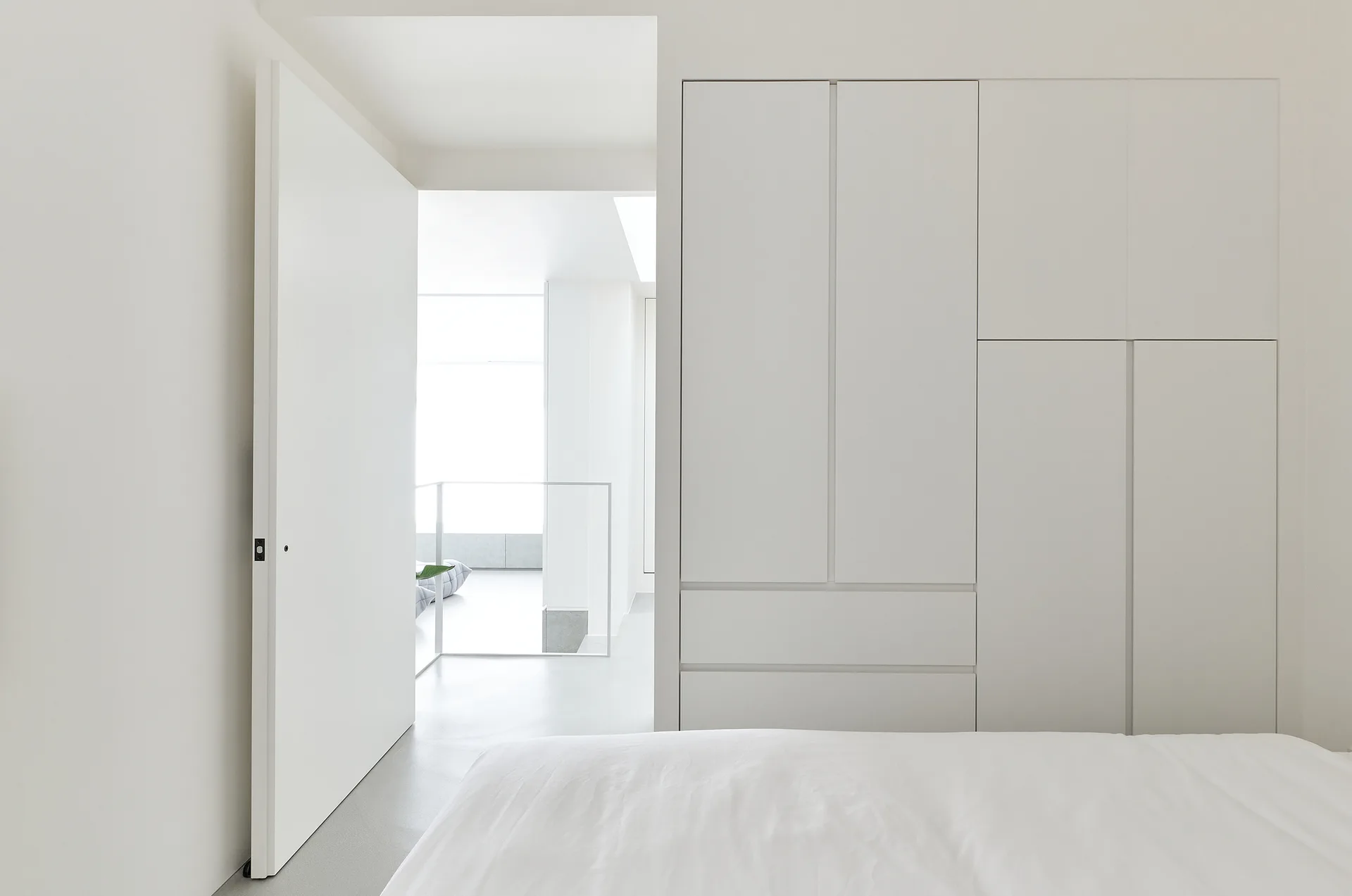 A minimalist white bedroom features integrated flat-panel wardrobe cabinets, a bed with white sheets, and an open door looking toward a staircase.
