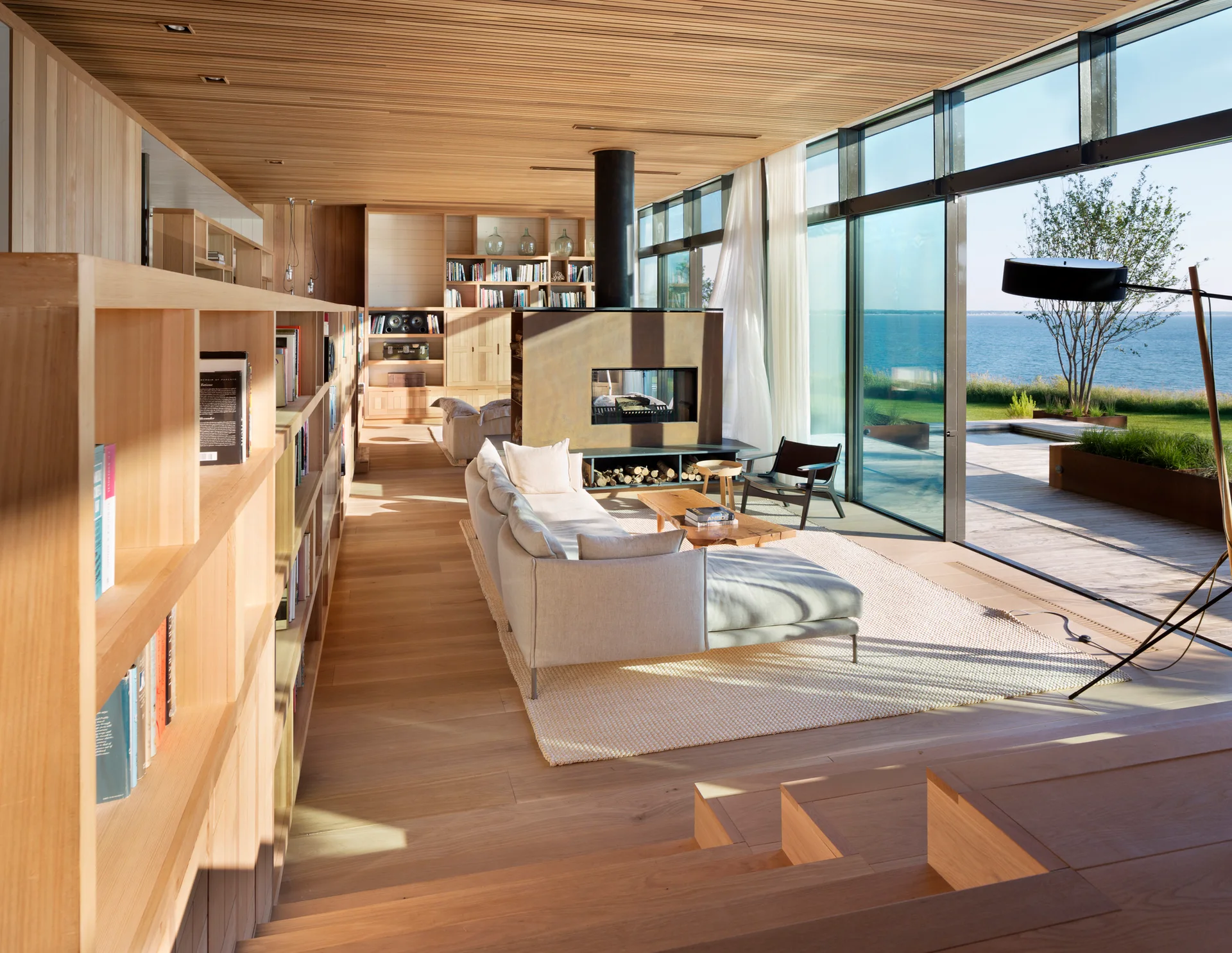 A modern coastal living room features a wood slatted ceiling, a freestanding fireplace, a beige sofa, and glass sliding doors looking out to the ocean.