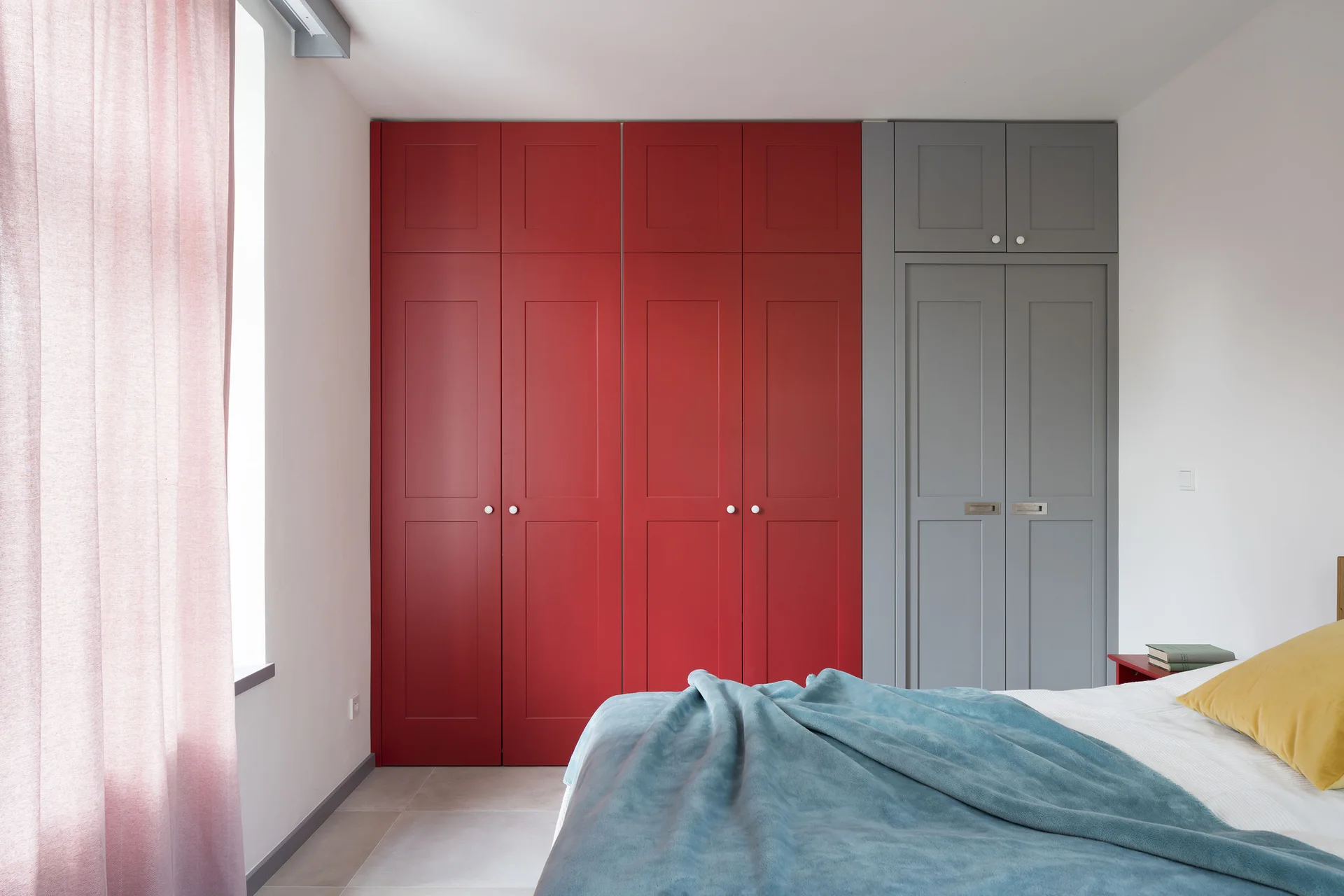 A modern bedroom with a large built-in wardrobe featuring four red shaker-style doors and two grey doors next to a bed with blue and yellow linens.